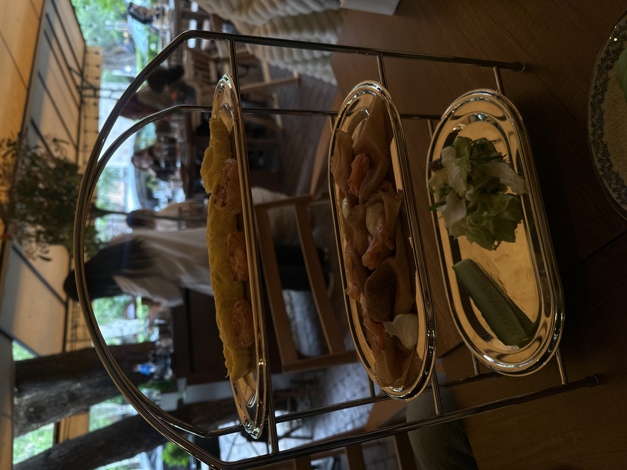 The image depicts a three-tiered food stand in what appears to be a restaurant in Yerevan, Armenia. The stand is made of a shiny metal, and each tier holds an oblong-shaped silver plate.
The first tier contains a long, rectangular food item that appears to be breaded and fried. The second tier holds crepes filled with salmon, and a white sauce. The third tier has a small pile of lettuce and a half of a cucumber.
In the background, there are several people sitting at tables, and there is a glimpse of greenery outside the restaurant. The lighting is somewhat dim, suggesting that it is either evening or the restaurant is dimly lit. The perspective is slightly tilted, giving a sense that the photo was taken at an angle.