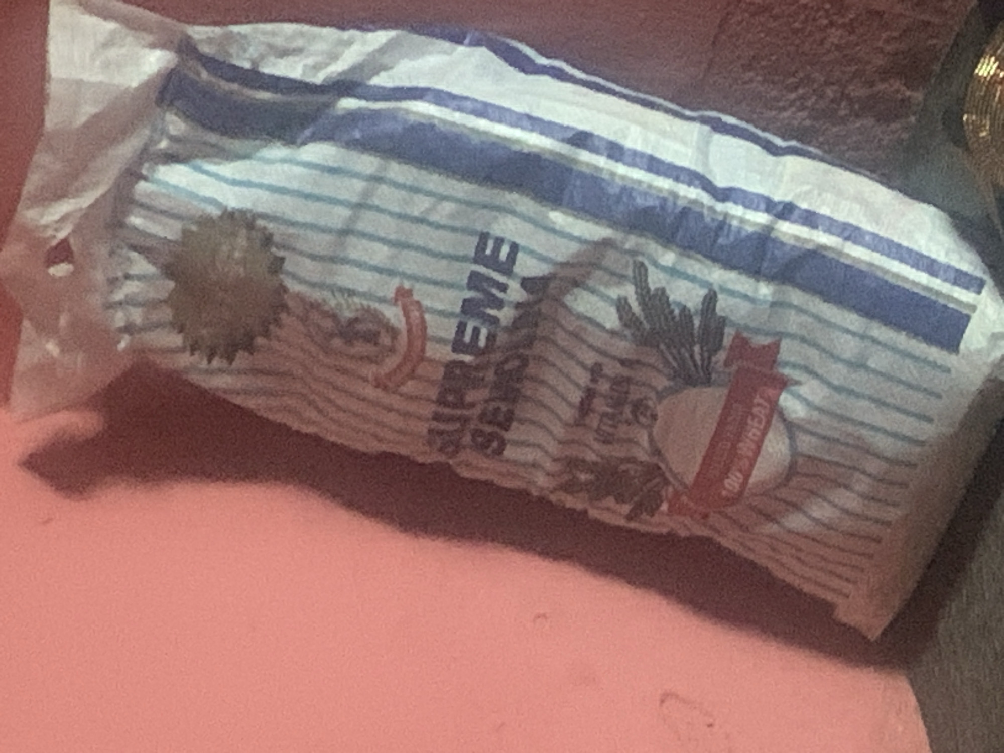 A tightly packed, vertically oriented, medium-shot image shows a bag of "SUPREME SEMOLINA" leaning against a pink wall. The bag is made of white plastic with thin blue vertical stripes and features a circular, spiky graphic at the top. Below this, text reads "SUPREME SEMOLINA," and further down, a red ribbon-like banner displays "100% WHEAT". The bag appears to be full, suggesting it is either a new purchase or has been recently filled. The lighting in the image is dim, casting a noticeable shadow of the bag on the wall to its left, indicating a light source from the right. The background consists of the pink wall and a darker, textured surface at the bottom, possibly a floor or table. The image is underexposed, with some blurriness, particularly on the right side. The scene is domestic, likely within a home in Jos, Nigeria, based on the provided context.