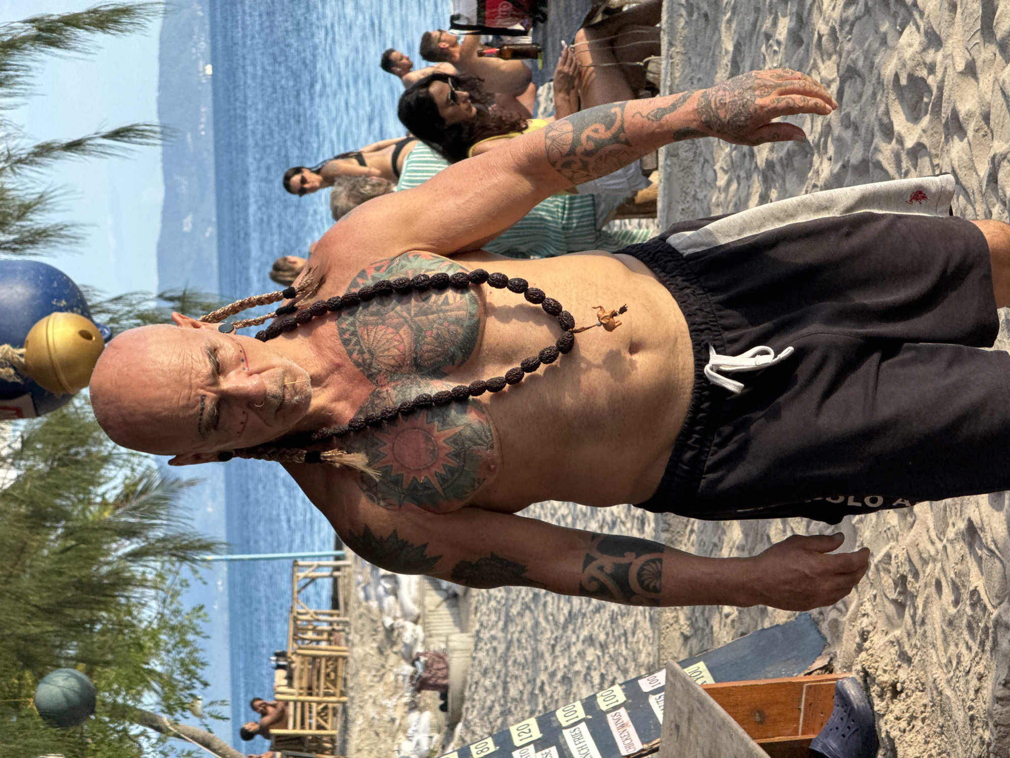 A man with tattoos on his chest and arms, a bald head, and braided hair extensions stands on a sandy beach. He is wearing black shorts with a white drawstring and a gray stripe on the side. Around his neck, he wears a beaded necklace with a small elephant pendant. Behind him, the ocean is visible, with a few people sitting at tables in the background. A price board is also visible to the left of the man. The scene appears to be located at a beachside establishment in Ko Phangan, Thailand.