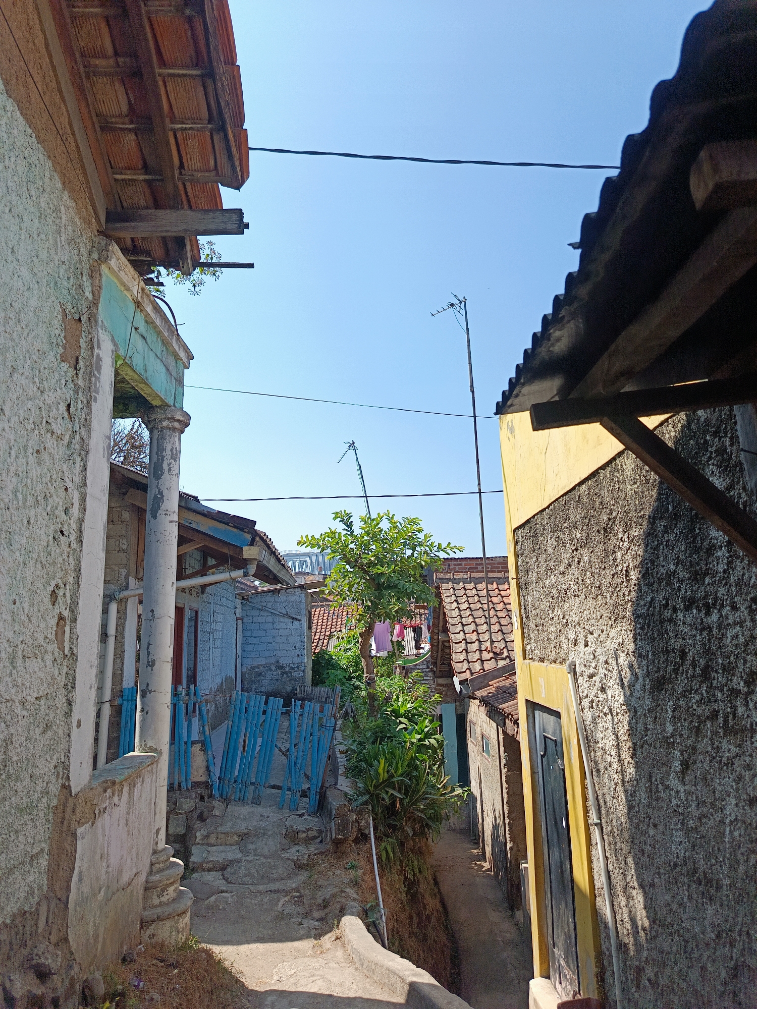 The image depicts a narrow alleyway in the city of Margajaya, Indonesia, bordered by two buildings. The building on the left has a weathered, light-colored textured facade with a partially visible blue-green trim and a large, peeling white pillar. The building on the right has a rough, grey textured facade with a bright yellow door frame and a dark-colored door.
The alley is paved with a dirt path that leads uphill and is uneven, with some stone steps visible. A small blue wooden fence is present on the left side of the path. Green foliage and trees with visible laundry hanging on a line can be seen further down the alley. The sky is clear and blue, with power lines crisscrossing the upper portion of the frame. The architecture suggests a residential area, likely with older construction. The lighting indicates it is daytime, with distinct shadows cast by the buildings.