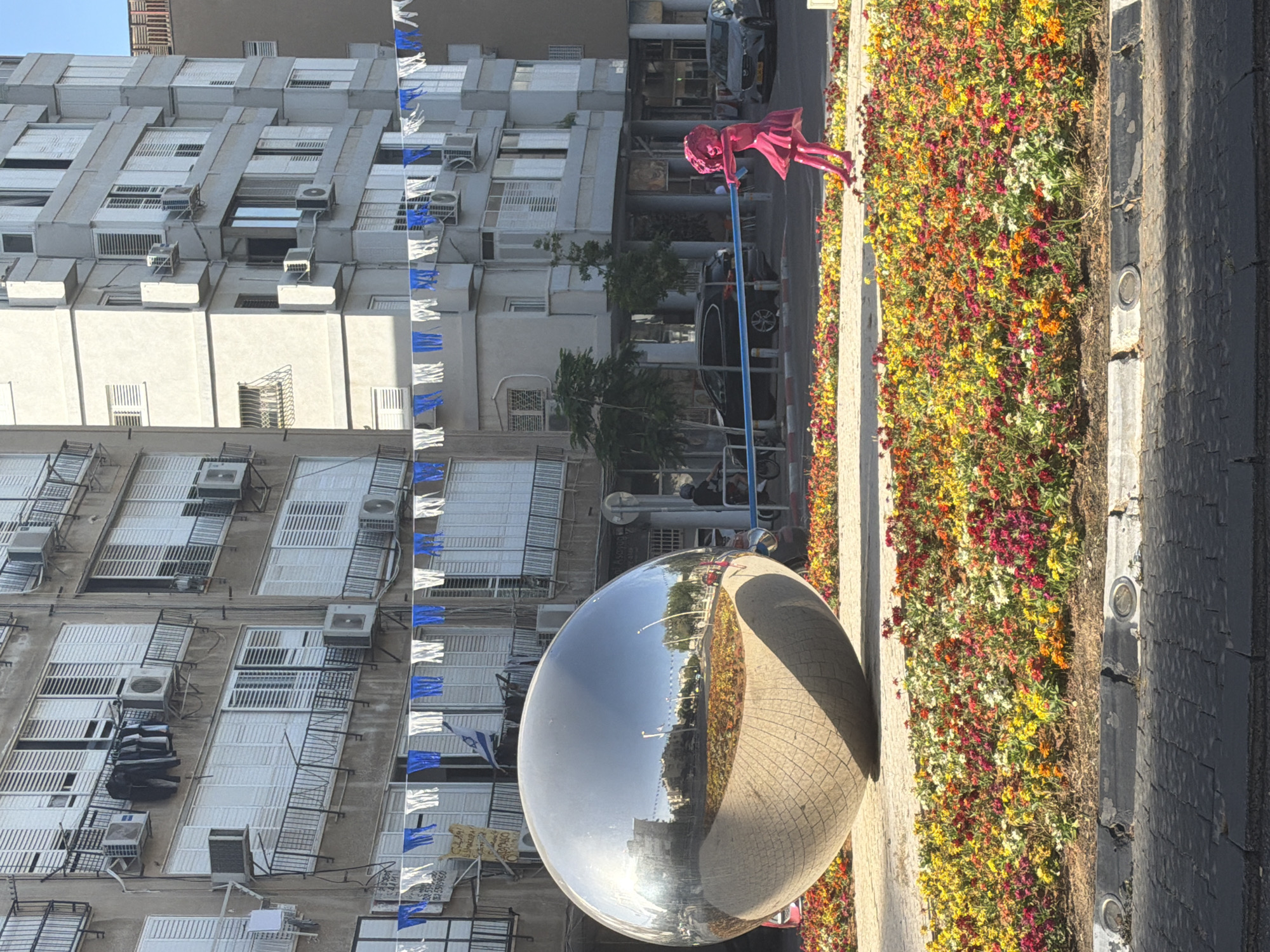 A street scene in Bat Yam, Israel, features a large, mirrored sphere reflecting the surrounding urban environment. The sphere sits near a flowerbed of red, yellow, and white flowers along the sidewalk. Buildings with multiple stories and rows of windows, some with air conditioning units and white metal grates, line the street. Blue and white pennants, possibly related to Israeli independence day or a similar celebration, hang from the buildings. In the upper right, a pink statue of a girl on a pole adds a pop of color. Cars are parked along the street. The sky is clear.