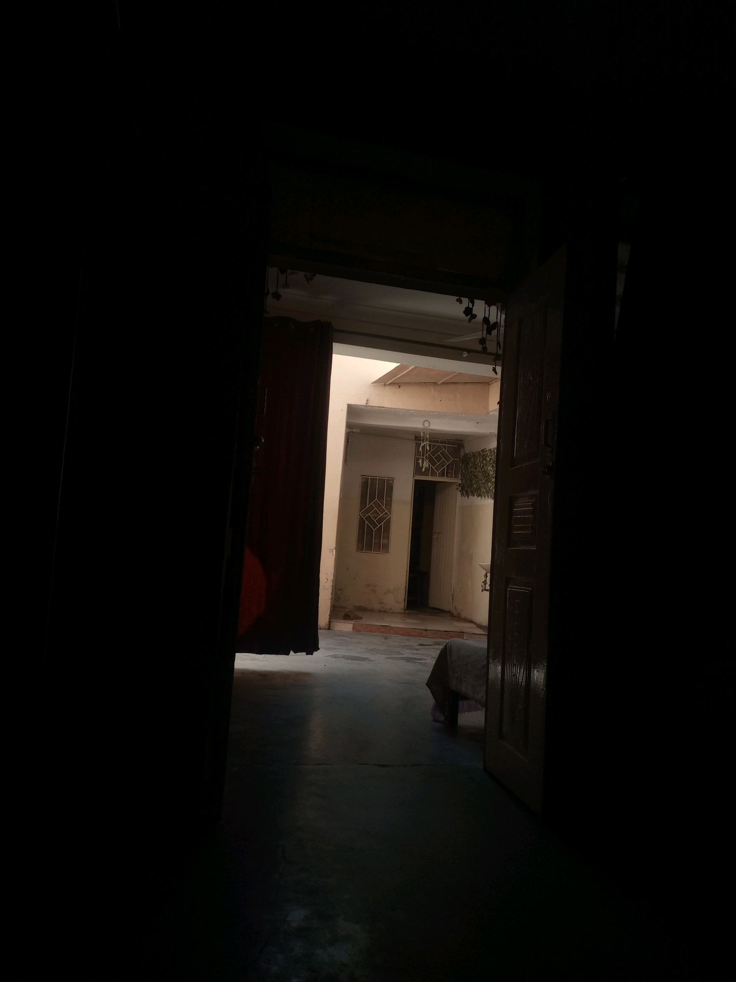 This is an interior shot looking through an open doorway towards a courtyard. The scene is dimly lit, with strong contrasts between light and shadow. On the left, a dark curtain hangs. On the right, a wooden door stands ajar, revealing more of the courtyard beyond. In the courtyard, there is a table covered with a light-colored cloth. The walls of the courtyard are painted a light color, and there are windows with metal grates visible. In the distance, there appears to be another doorway or opening. The floor is a smooth, dark surface. The overall impression is one of a quiet, possibly abandoned, domestic space. The image does not provide specific clues to pinpoint the location within Hasan Abdal, Pakistan, beyond being an interior courtyard.