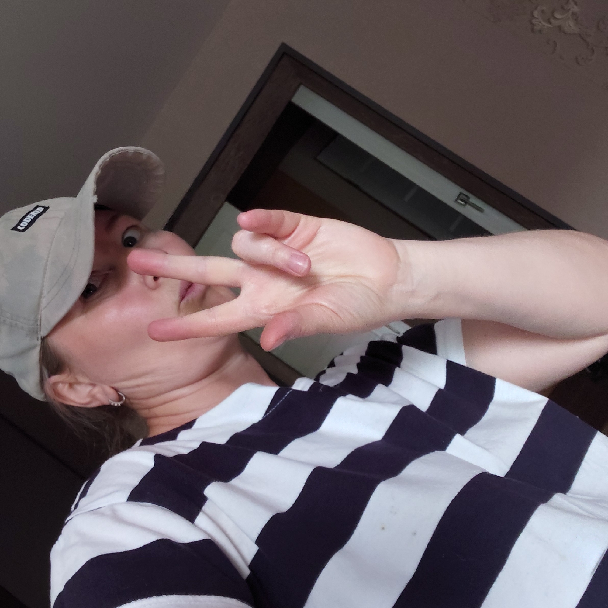 A close-up shot shows a person wearing a light-colored baseball cap and a navy and white striped shirt. They are holding up two fingers in a peace sign gesture. The background is blurred, but appears to be an indoor setting with a dark-colored door frame and a brown wall. The person's face is partially visible, with their eyes looking directly at the camera. The lighting is soft, and there are no discernible landmarks or specific architectural features that would indicate a location within Novosibirsk, Russia.