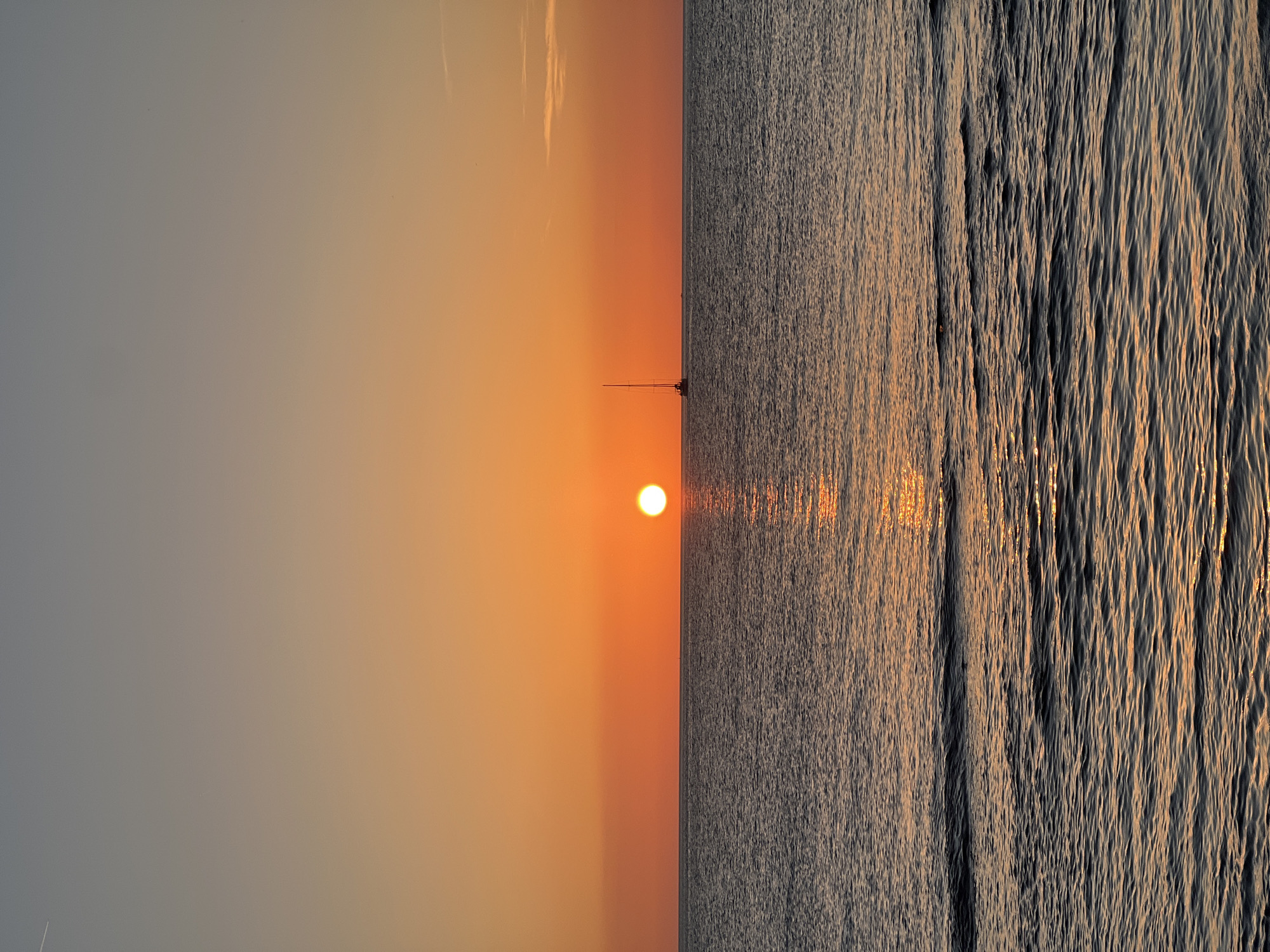 This image depicts a serene seascape at sunrise, likely near Palermo, Italy. The sun, a bright orb casting a warm glow, is just beginning to ascend, painting the sky in gradients of soft orange and pale gray. The horizon is sharply defined, separating the tranquil sky from the textured water.
A lone sailboat with a tall mast stands silhouetted against the vibrant sky, its form a subtle yet prominent detail in the vast expanse. The water is calm but not entirely still, with gentle ripples catching the sunlight and creating a shimmering effect. The waves are small and regular, suggesting a peaceful morning on the sea.
The overall mood of the image is one of tranquility and natural beauty. The soft light and gentle movement of the water evoke a sense of calm and peace. The absence of any human figures contributes to the feeling of solitude and vastness. The scene appears to be an open body of water, possibly the Mediterranean Sea, given the Palermo, Italy context. The time of day is clearly early morning, as indicated by the low position of the sun and the soft, diffused light. There are no visible clouds of significance, suggesting clear weather. No text is visible in the image.