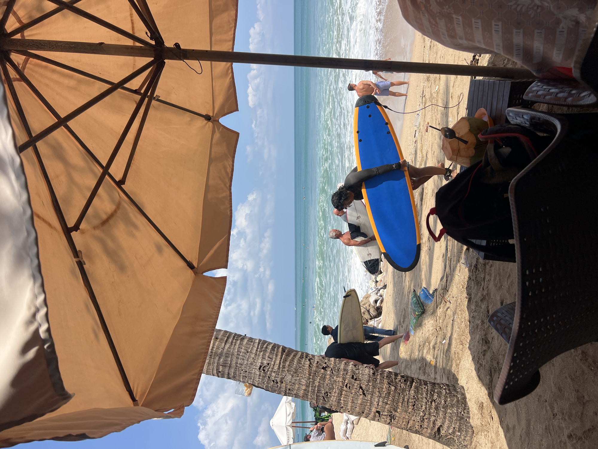 In Kuta, Indonesia, on a sunny day with a bright blue sky and scattered clouds, a beach scene unfolds under a large, tan umbrella. The foreground shows the edge of a beach chair and a table with a coconut drink and a bag. Beyond, a sandy beach is dotted with a palm tree, surfboards, and several people. Some individuals are carrying surfboards, suggesting they are either preparing to surf or have just finished. The ocean is visible in the background, with waves rolling onto the shore and a few surfers in the water. A white umbrella and some lounge chairs are set up further down the beach, indicating a leisure area. The overall atmosphere appears relaxed and recreational, typical of a beach resort.