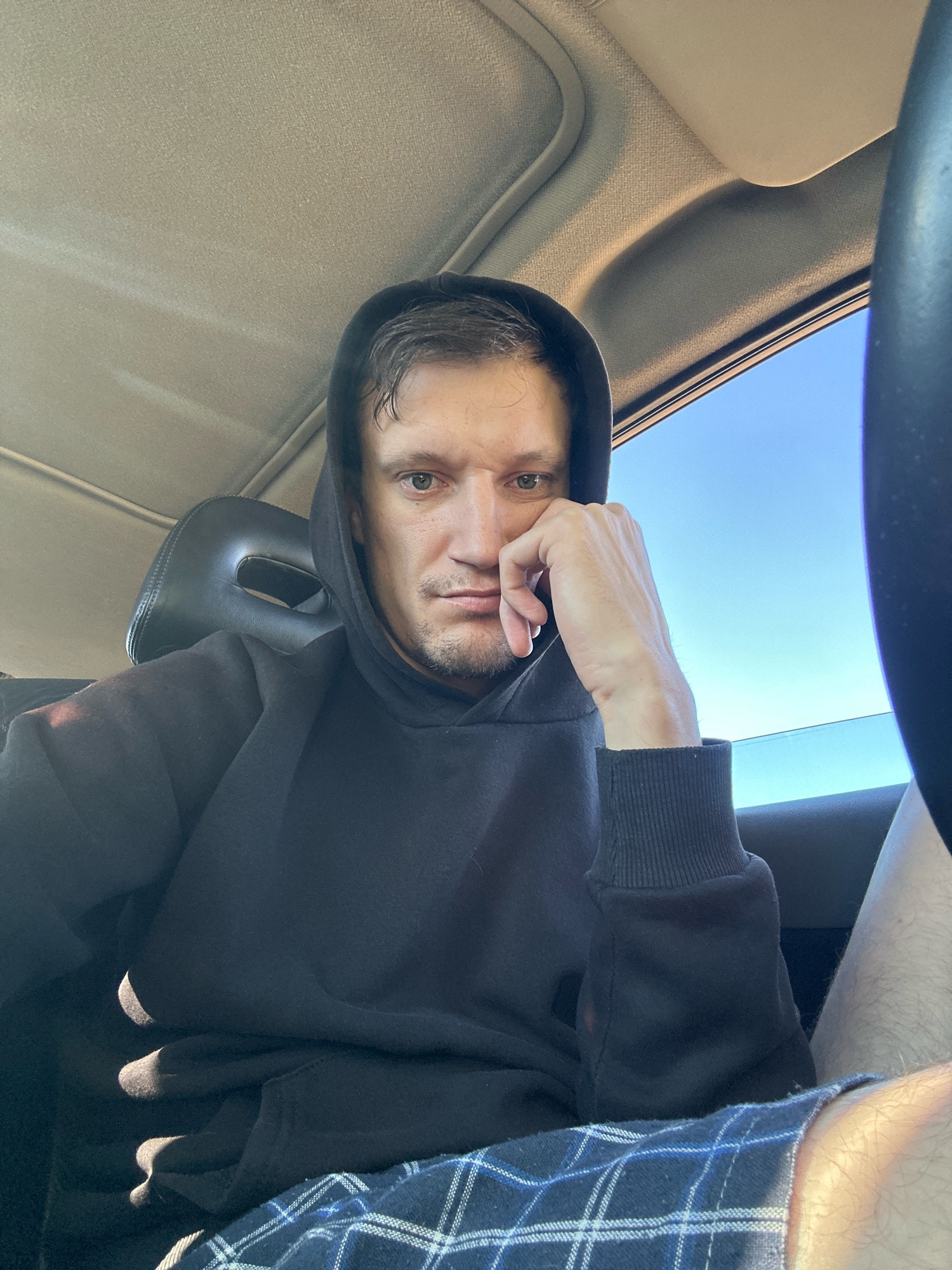 A man is sitting in the driver's seat of a car, looking at the camera. He is wearing a black hoodie and plaid shorts. His right hand is resting on his chin. The car's interior is visible, including the headrest and the ceiling. Sunlight streams in through the window, illuminating the scene. The background shows a clear blue sky. The image was taken in Makiv, Ukraine.