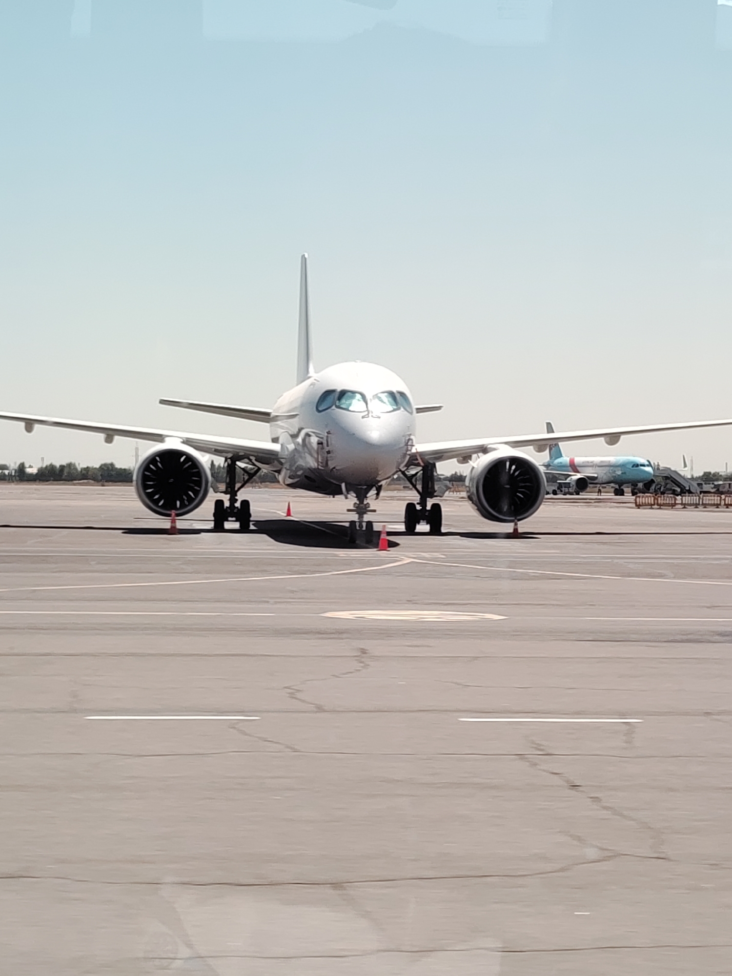 A wide-angle, eye-level, frontal view captures a large, white passenger airplane parked on the tarmac of an airport. The aircraft is the primary subject, dominating the center of the frame with its nose pointed directly at the camera. Its wings extend outwards, revealing two large engines beneath them. The undercarriage is visible, with landing gear supporting the plane. Orange traffic cones are placed strategically around the aircraft.
In the background, a second, smaller airplane, primarily blue and white, is visible on another section of the tarmac, parked with a set of stairs leading to its cabin. Additional airport infrastructure, including fences and distant buildings, can be seen. The scene is set against a clear, pale blue sky. The ground is a grey asphalt surface marked with white lines, indicative of a runway or taxiway. The overall environment suggests a sunny day at an airport.
The airplane's registration is partially visible as UK-38005, and the airline's logo, "Uzbekistan Airways," is present on the tail fin of the blue and white aircraft in the background. The scene is located at Tashkent International Airport, officially known as Islam Karimov Tashkent International Airport (IATA: TAS, ICAO: UTTT), in Tashkent, Uzbekistan. No people are clearly discernible within the frame.