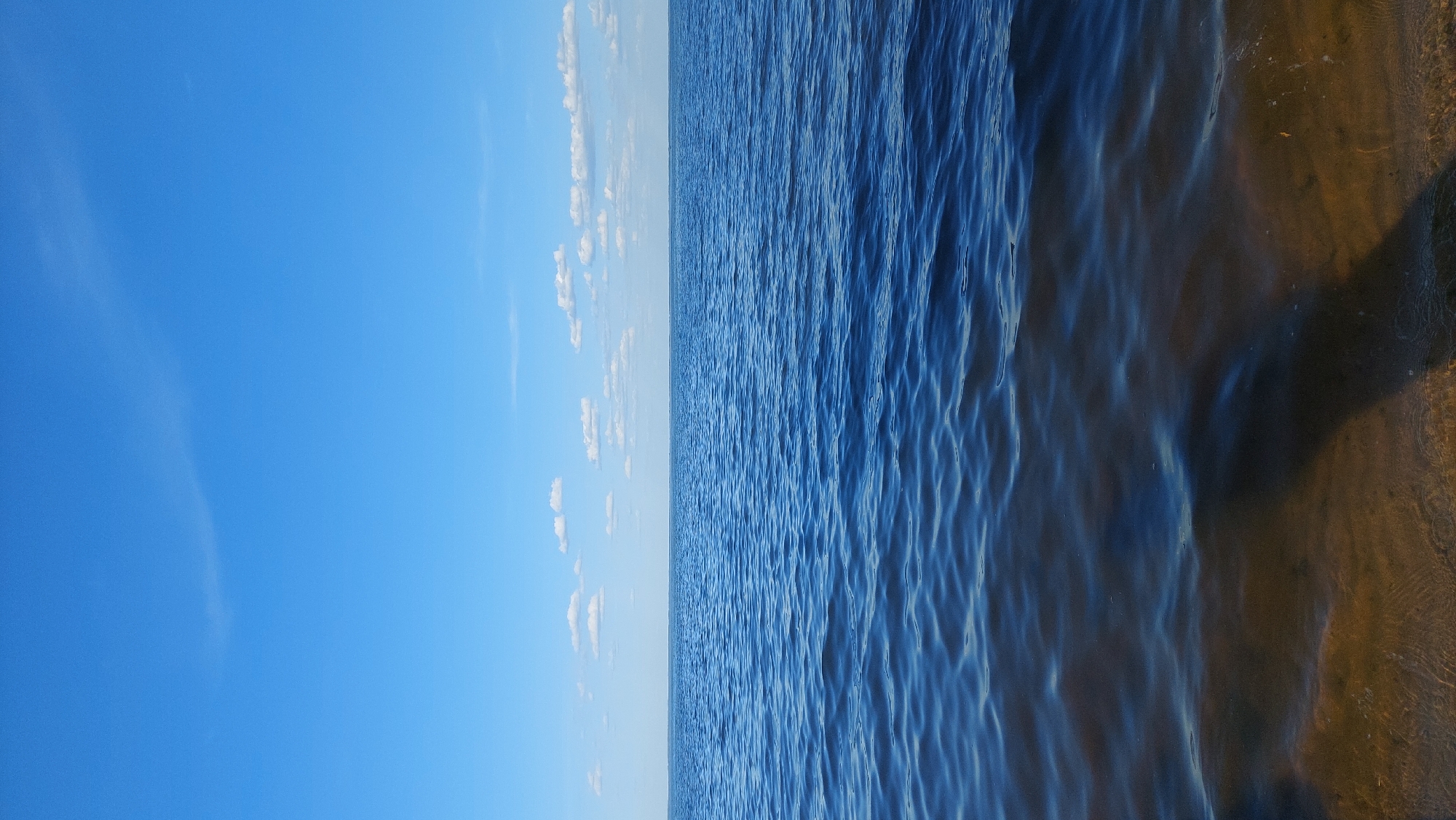 The image depicts a serene natural scene in Kozarovychi, Ukraine. The horizon line divides the frame, with the upper portion dominated by a vast, clear blue sky. A few scattered, wispy white clouds drift lazily across the sky. Below the sky lies a wide expanse of deep blue water, likely a lake or river, with gentle ripples disturbing its surface. The water appears calm and reflects the blue of the sky. The foreground of the image shows the shallow edge of the water, where the sandy or muddy bottom is visible through the translucent water. A dark shadow, possibly cast by a person or an object not fully within the frame, stretches across the sandy bottom. The overall impression is one of tranquility and natural beauty.