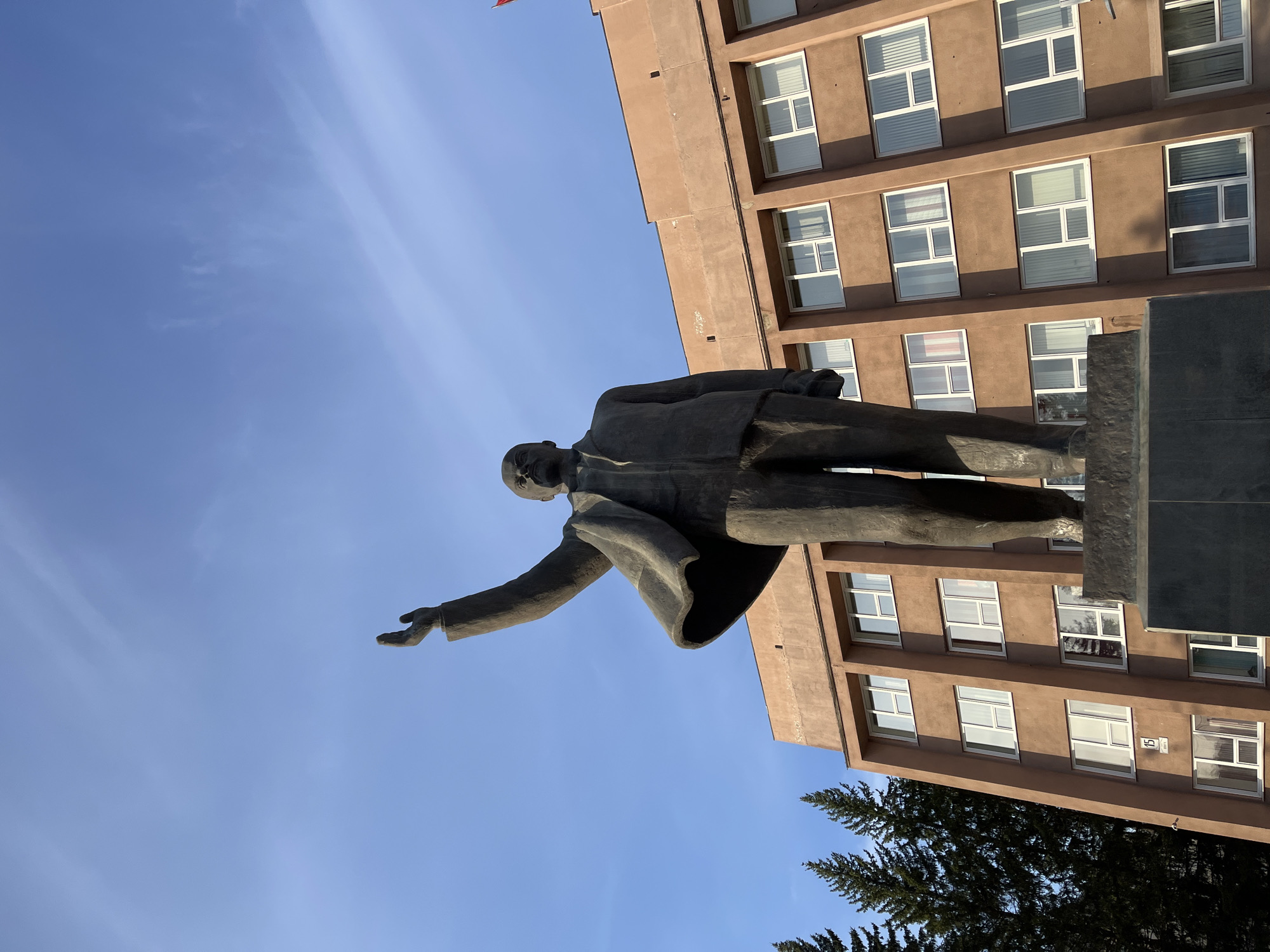 The image depicts a statue of Vladimir Lenin, likely located in Zelenogorsk, Russia, given the architectural style of the building in the background, which is characteristic of Soviet-era construction. The statue is positioned in front of a multi-story building with a brown facade and numerous windows. Lenin is depicted in a full-body pose, gesturing forward with his right arm outstretched. The sky is visible in the background, and part of a tree is visible.