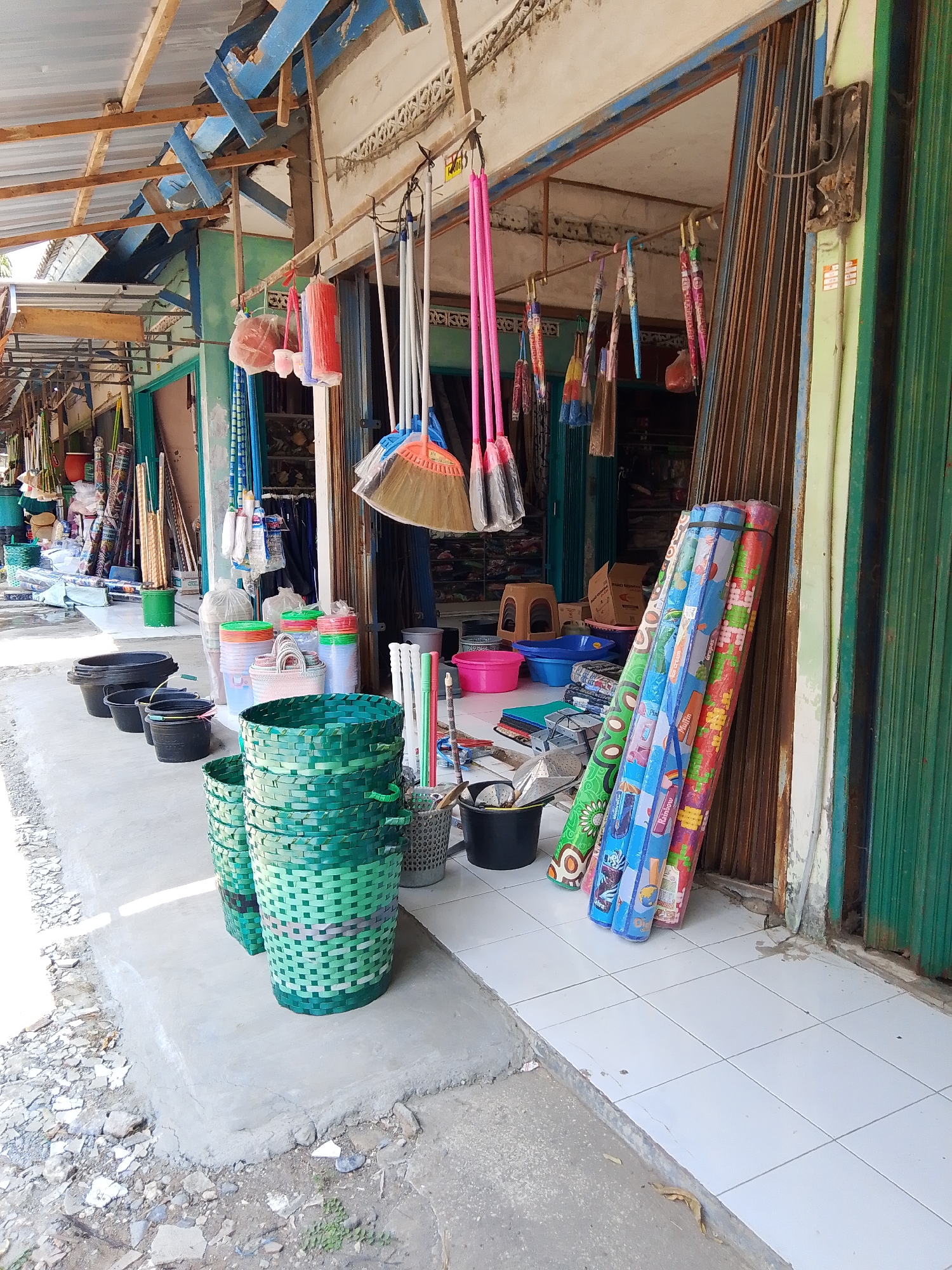 The image depicts the exterior of a shop in Kedondong, Indonesia, likely a market stall or small convenience store. The storefront is open, revealing a variety of household items for sale. On the right, a stack of rolled-up mats with colorful patterns leans against a corrugated metal gate. To the left of the mats, various cleaning tools such as brooms with brightly colored handles (pink, blue, and orange) are displayed hanging from a horizontal bar. Further to the left, several green woven baskets are stacked, with smaller black buckets and tubs lined up in front of them. In the background, more items are visible, including stacks of plastic containers and what appear to be more woven baskets. The shop has a weathered wooden and metal roof structure. The ground in front of the shop is paved with light-colored tiles and a concrete walkway, with rougher ground and debris on the left side. There are no people visible in the image.