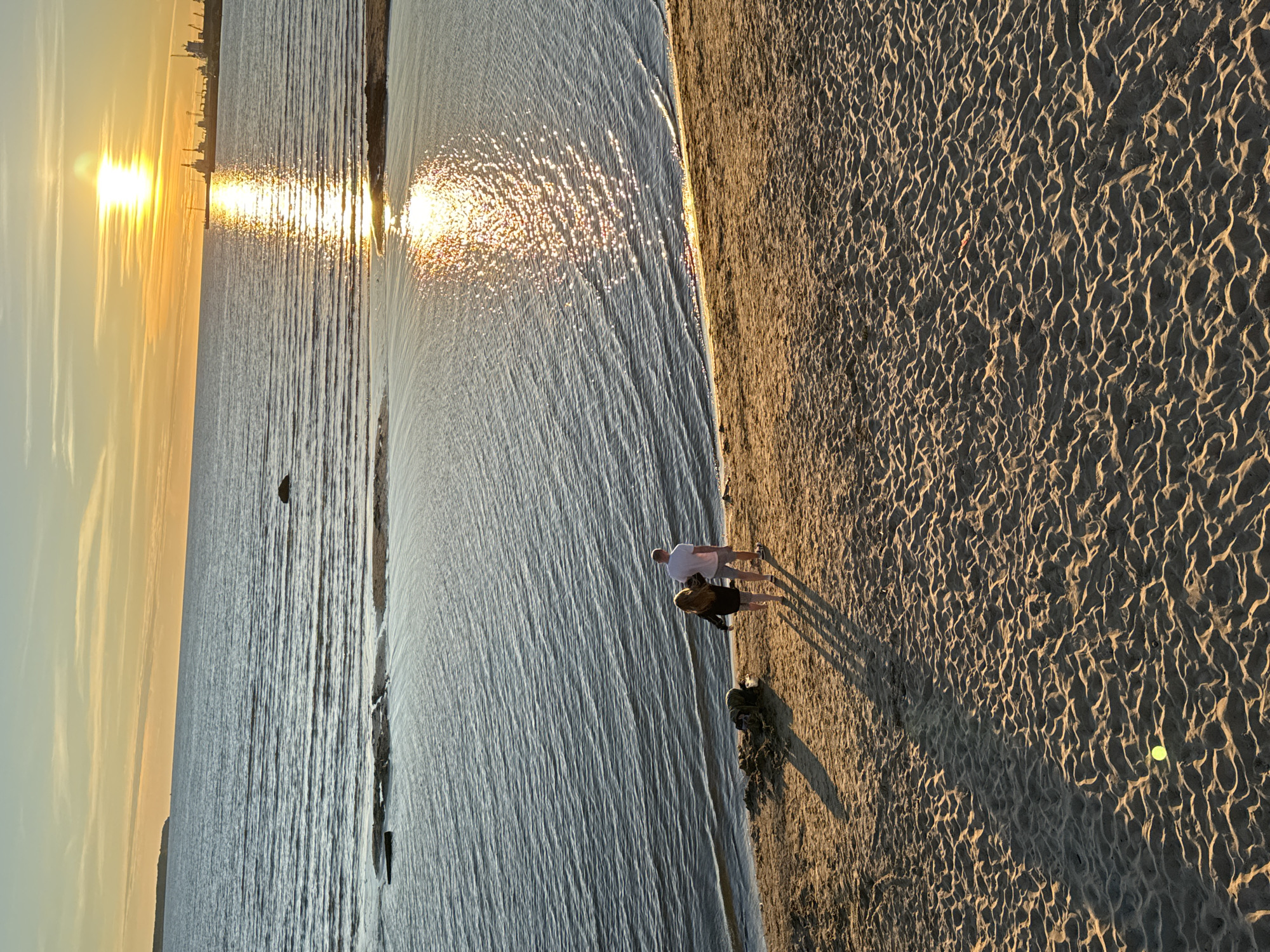 This is a high-angle, long shot of a sandy beach area at sunset, located in the Pirita district of Tallinn, Estonia. Two individuals, a man and a woman, are walking away from the camera towards the water. The man is wearing a white t-shirt and grey shorts, while the woman is wearing a dark top and shorts. They are casting long shadows on the sand. A small pile of dark-colored items, possibly bags or clothing, is located to their left on the sand. The water in front of them is calm with ripples, reflecting the golden light of the setting sun. In the distance, across the water, a port with cranes and ships is visible, silhouetted against the bright sky. The sky is a gradient of pale blue and orange, with the sun a brilliant white orb low on the horizon. The sand in the foreground is textured with ripples and shadows, and a single yellow tennis ball is visible on the sand.