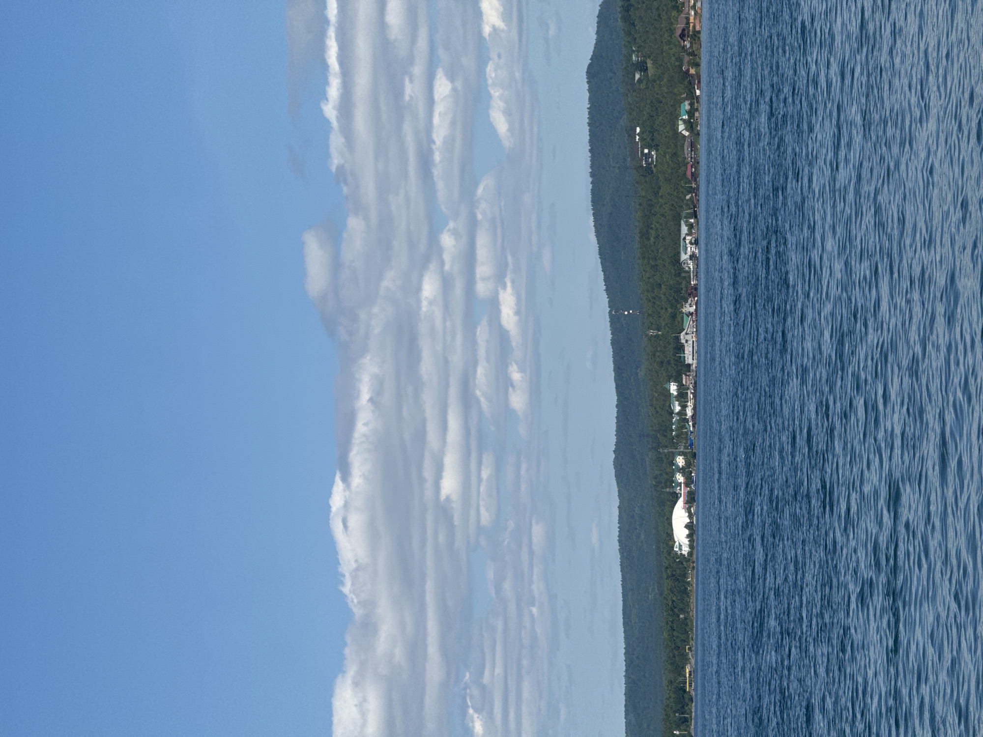 A scenic view of the Listvyanka settlement on the shore of Lake Baikal, Russia, is captured under a bright blue sky dotted with fluffy white clouds. The foreground is dominated by the deep blue waters of Lake Baikal, with gentle ripples suggesting a calm day. In the middle ground, the settlement of Listvyanka stretches along the coastline, nestled at the foot of a densely forested hill. Several buildings are visible, including a large, white domed structure, likely a tourist facility or museum. Houses with red and brown roofs are scattered along the shore. A prominent transmission tower can be seen on the forested hillside. The background features rolling, tree-covered hills that form the natural landscape surrounding Listvyanka.
