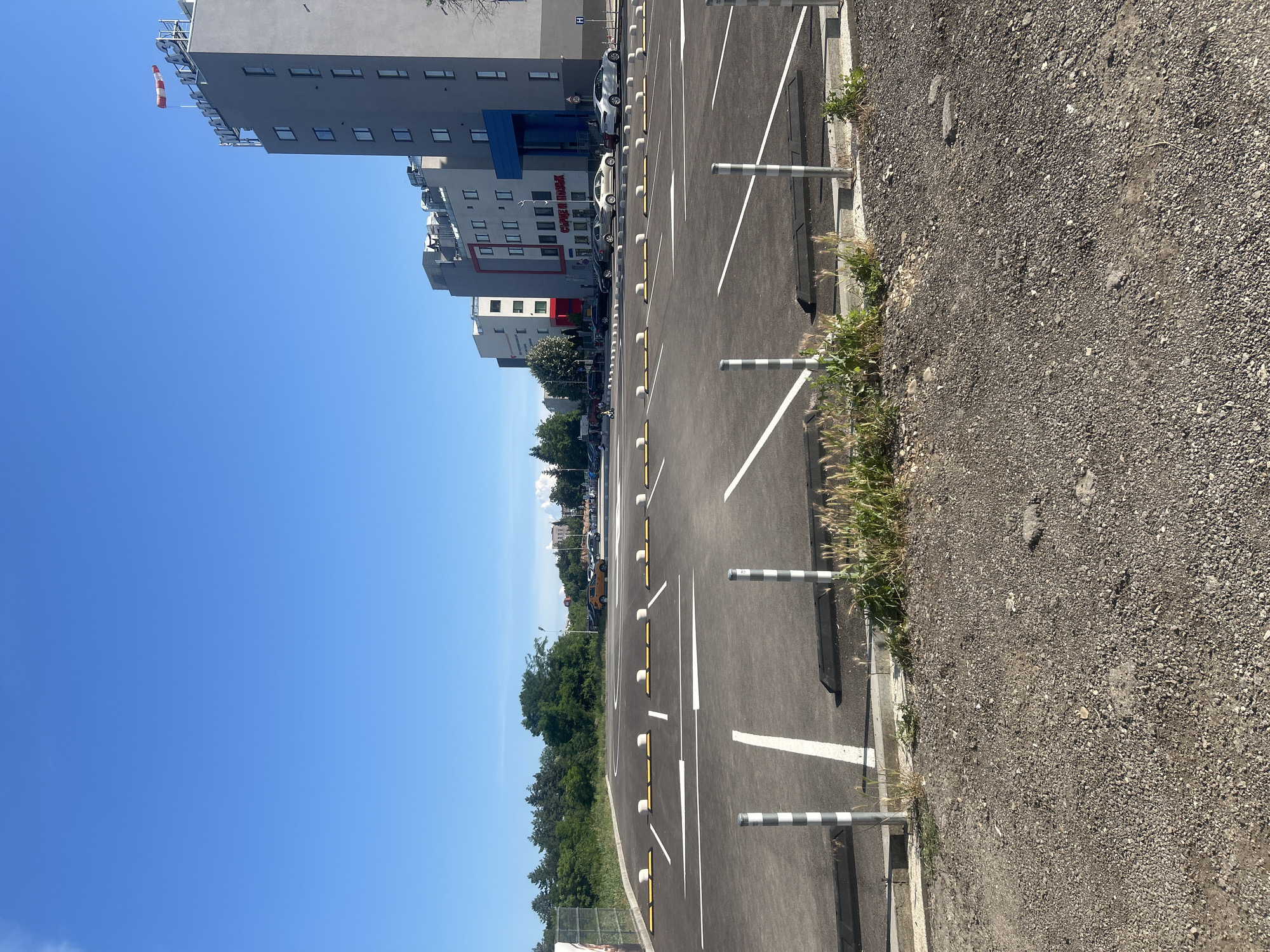 The image shows an empty outdoor parking lot with newly painted white lines and yellow and white wheel stops. In the background, there is a modern multi-story building complex with several cars parked in front. The building has signage that appears to be in Bulgarian. A windsock is visible at the top of one of the buildings. The sky is clear and blue. The foreground of the image shows a gravelly area with some weeds growing. The overall scene suggests an urban environment, likely in Pleven, Bulgaria, given the architectural style and Cyrillic signage.