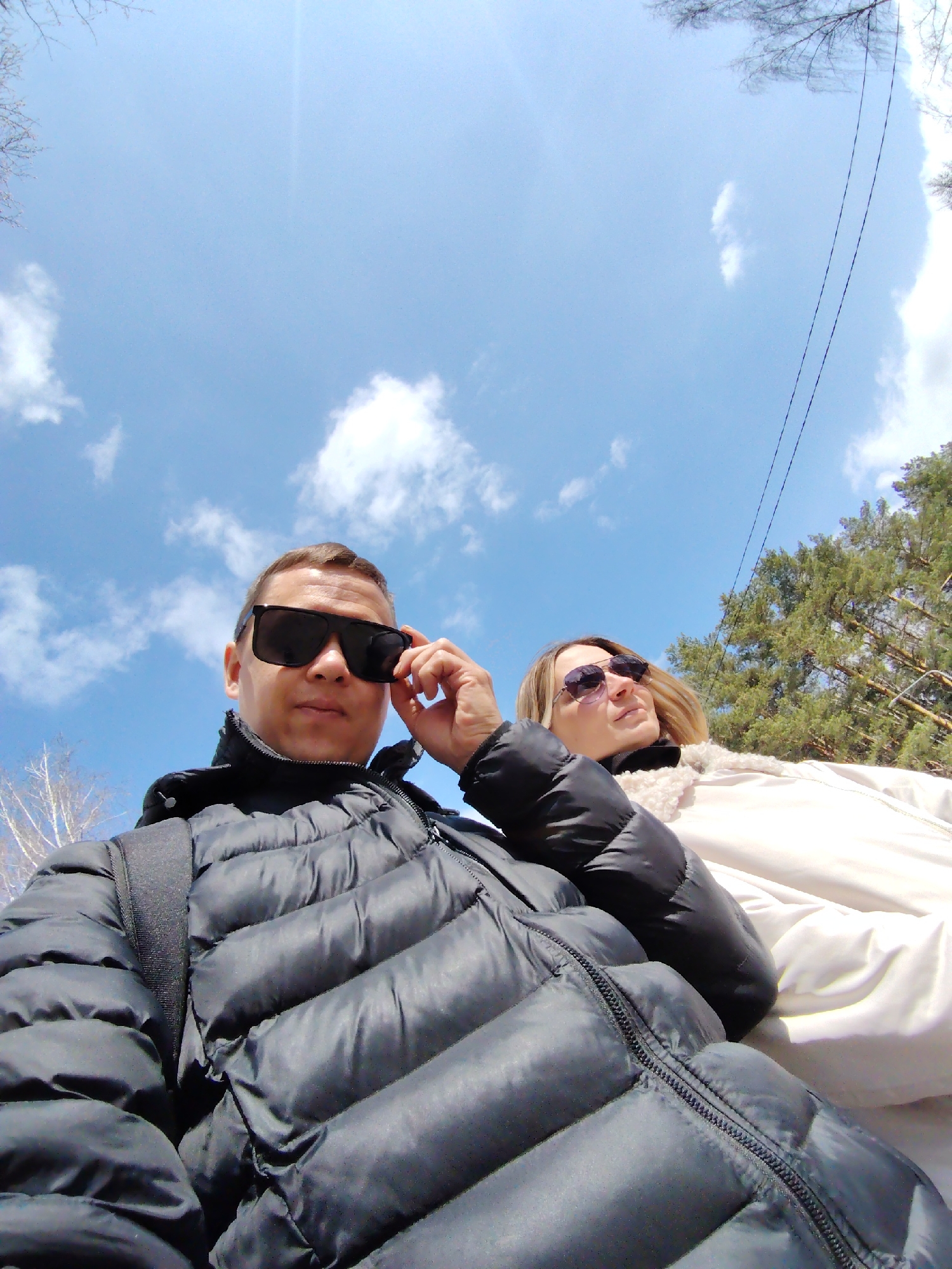A low-angle shot captures two individuals outdoors in Chaikovsky, Russia. A man, sporting dark sunglasses, is dressed in a puffy black jacket and has a black backpack slung over his shoulder. He's adjusting his glasses with his right hand. Beside him, a woman wears tinted sunglasses and a cream-colored jacket with a fluffy collar. The background features a bright blue sky dotted with white clouds, and trees are visible to the right. Electrical wires are also present against the sky. Bare tree branches are seen in the upper left corner.