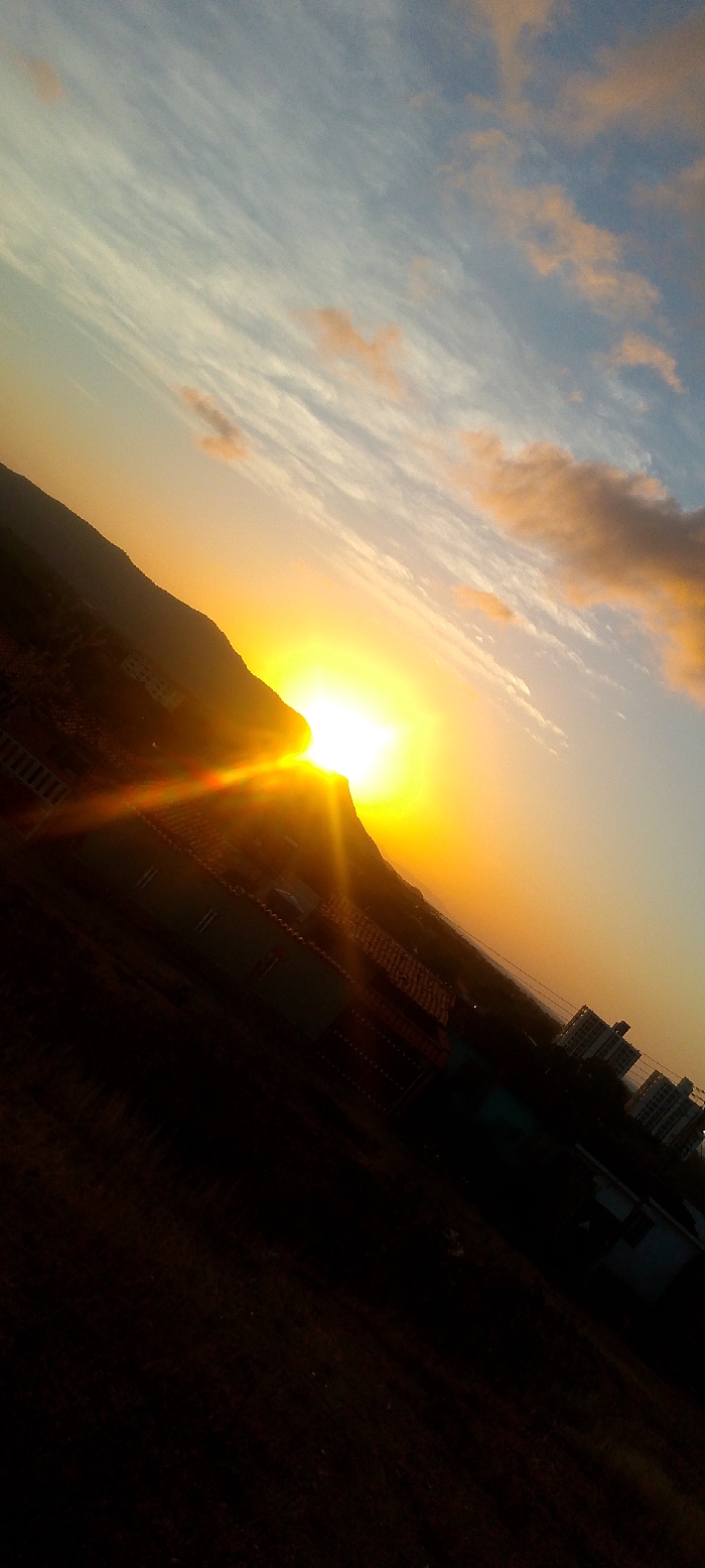 The image displays a panoramic view during either sunrise or sunset over the city of Juan Griego, Venezuela. The scene is captured with a diagonal tilt, approximately 30-45 degrees clockwise. A bright, intense yellow-orange sun is positioned behind a large, dark, silhouetted hill or mountain, which dominates the left side of the frame and partially obscures the sun. Significant lens flare emanates from the sun, casting strong light rays downwards and to the left. The sky transitions from a light blue with scattered wispy white clouds and some orange-tinted cumulus clouds in the upper right to brilliant yellow and orange hues near the sun. In the midground, below the primary landmass, a building with a reddish-brown tiled roof is partially visible, appearing dark due to backlighting. Further to the right, a cluster of several multi-story, light-colored buildings are silhouetted against the sky. The lower foreground is heavily underexposed, appearing as dark, indistinct terrain or sparse vegetation. No individuals, discernible actions, or interactions are visible within the frame.