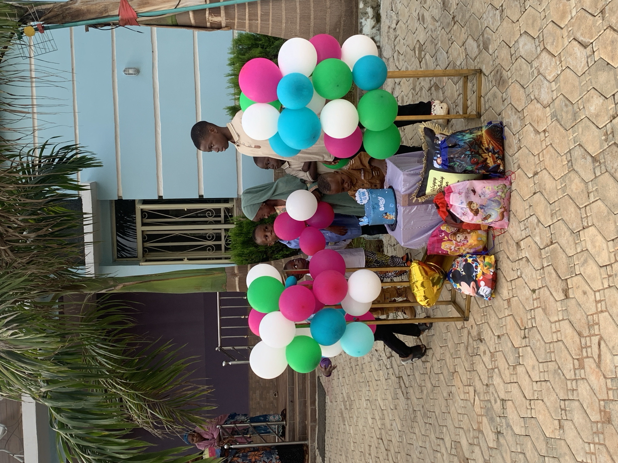 This image captures an outdoor birthday celebration in the city of Jos, Nigeria. The scene is set on a light brown, intricately paved area resembling interlocking hexagons and rectangles, with residential buildings and lush vegetation in the background.
In the foreground, a central arrangement features a small, light purple-draped table holding a blue, round birthday cake. The cake is decorated with white frosting and features a sign that reads "FAHEEM @ ONE" and "BLUEY" with additional text "Happy B'day". A young boy, presumed to be Faheem, wearing a brown shirt, is seated or kneeling in front of the table, looking down at the cake. Surrounding the cake table are various gift bags on the ground, depicting characters such as Mickey Mouse, Disney princesses (including Sofia the First), and Marvel superheroes. A "Happy Birthday" themed pillow is also visible. To the left of the table, a gold star-shaped balloon rests near the gifts.
Flanking the central celebration area are two tall arrangements of balloons. These clusters consist of spherical balloons in white, bright green, magenta, and light blue. Several children are present, gathered around these arrangements. To the right of the right balloon cluster, an older boy in a light beige collared shirt stands upright, looking towards the left. A young girl wearing a green hijab is visible behind the birthday boy, observing the cake. Other children, some partially obscured by the balloons, are also present, including a young boy in a blue shirt on the far left.
In the mid-ground, on the left, a purple-walled building with a white trim and a metal-railed balcony is visible. Two adult females are on this balcony; one is leaning on the railing, wearing a pink top and patterned skirt, while the other is further back, wearing a blue headscarf and a patterned dress. Steps lead up to this building. On the right, a light blue building with horizontal white stripes and a window secured with white bars is visible. A light fixture is mounted on its exterior wall.
The environment includes several large palm trees with long, green fronds, providing shade. Green shrubs are visible at the base of the blue building. The lighting is bright and even, indicating daylight. An antenna-like structure with a yellow circular component is visible attached to a tree trunk on the far right.