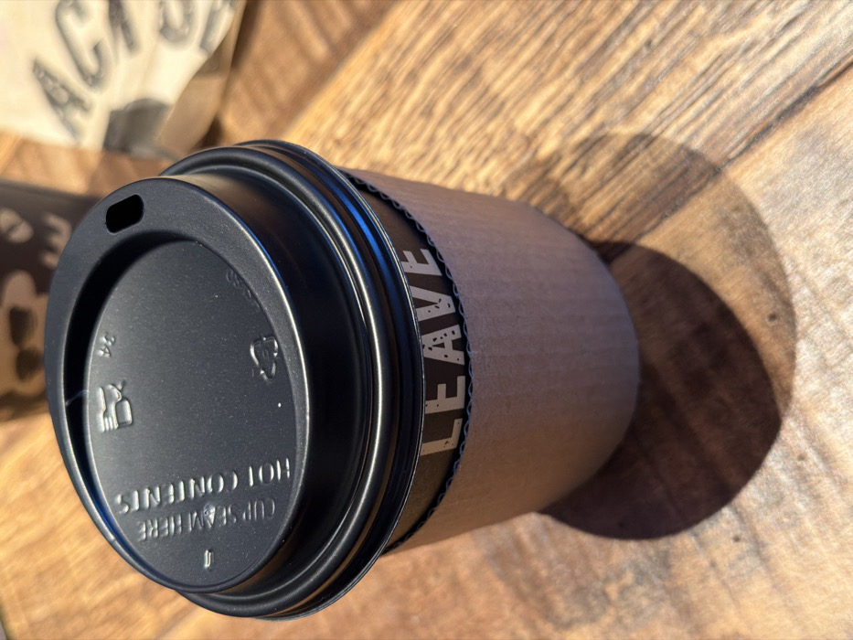 The image displays a disposable coffee cup with a black plastic lid and a corrugated cardboard sleeve, positioned on a light brown wooden table. The black lid features an oblong drinking opening, a raised rim, and embossed text stating "CUP SEAM HERE" and "HOT CONTENTS," alongside a partial recycling symbol and a utensil icon. The natural brown cardboard sleeve displays the word "LEAVE" in white, stippled capital letters against a dark background band. The wooden table surface exhibits distinct grain patterns, suggesting horizontal planks. Strong directional lighting from the upper left casts a clear, elongated shadow of the cup onto the table. In the blurred background, a second, dark-colored disposable cup with light patterns is partially visible, along with a crumpled brown paper item displaying indistinct light text. No individuals or specific actions are depicted. The scene is consistent with a commercial establishment such as a coffee shop or cafe located in Bretton, United Kingdom; however, a precise address within Bretton cannot be determined from the visual evidence.