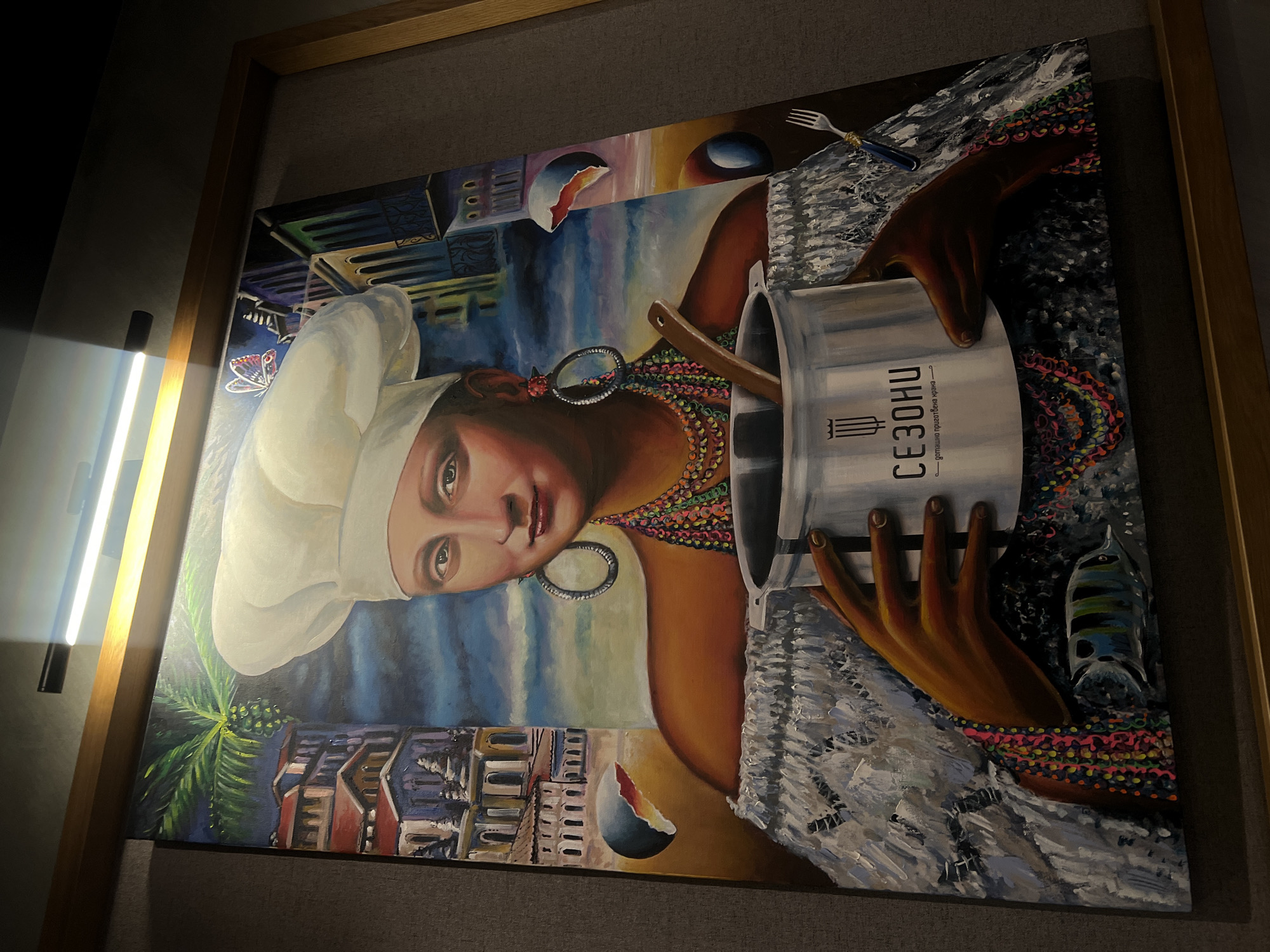 A framed painting hangs on a wall. The painting depicts a person with dark skin wearing a chef's hat, large hoop earrings, and a multi-colored beaded necklace. The person is holding a silver pot with the word "СЕЗОНИ" written vertically on it. A wooden utensil is visible inside the pot. A hand with brown skin is holding the pot. In the background, there is a cityscape with buildings and a blue sky. There is also a fork placed above the cityscape. The painting is in a brown wooden frame. A light source is visible on the left side of the image, creating a glare. I am unable to find out from the image if it is located in Sofia, Bulgaria.