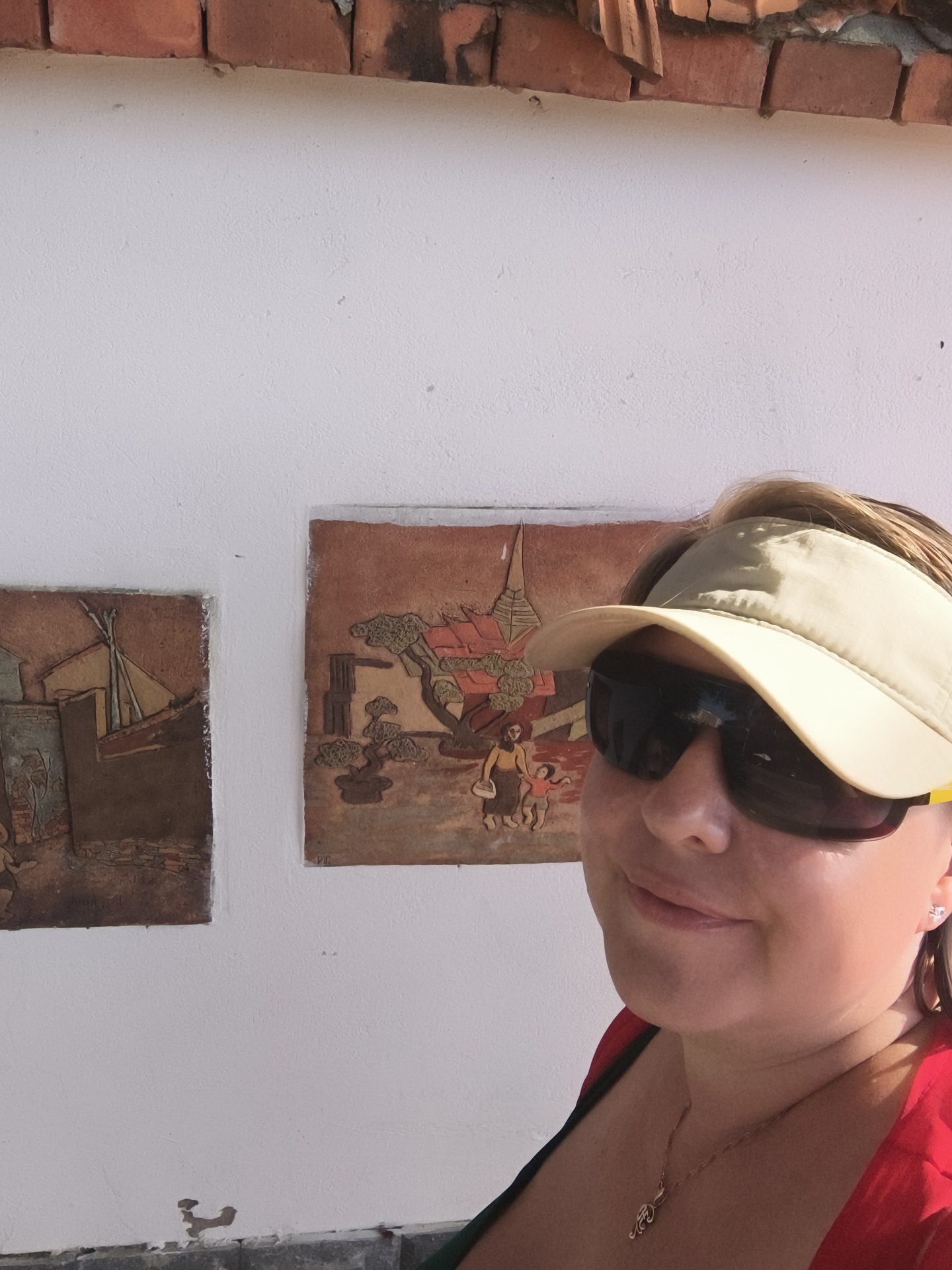 The image shows a woman with blonde hair wearing a tan visor and dark sunglasses. She is also wearing a red shirt and a gold necklace with a pendant. In the background, there are two framed artworks on a white wall. Above the white wall, there is a row of red bricks. The artworks appear to be relief carvings depicting village scenes. The artwork on the left shows a structure resembling a hut and some foliage. The artwork on the right shows a larger structure with a conical roof, trees, and two figures, one appearing to be an adult holding the hand of a child. The woman is positioned to the right of the artworks, and her face is close to the camera, with only her upper body visible. The overall lighting suggests an outdoor setting with bright sunlight. It is not possible to determine the exact location within Phu Thuy Ward, Vietnam from the image alone.