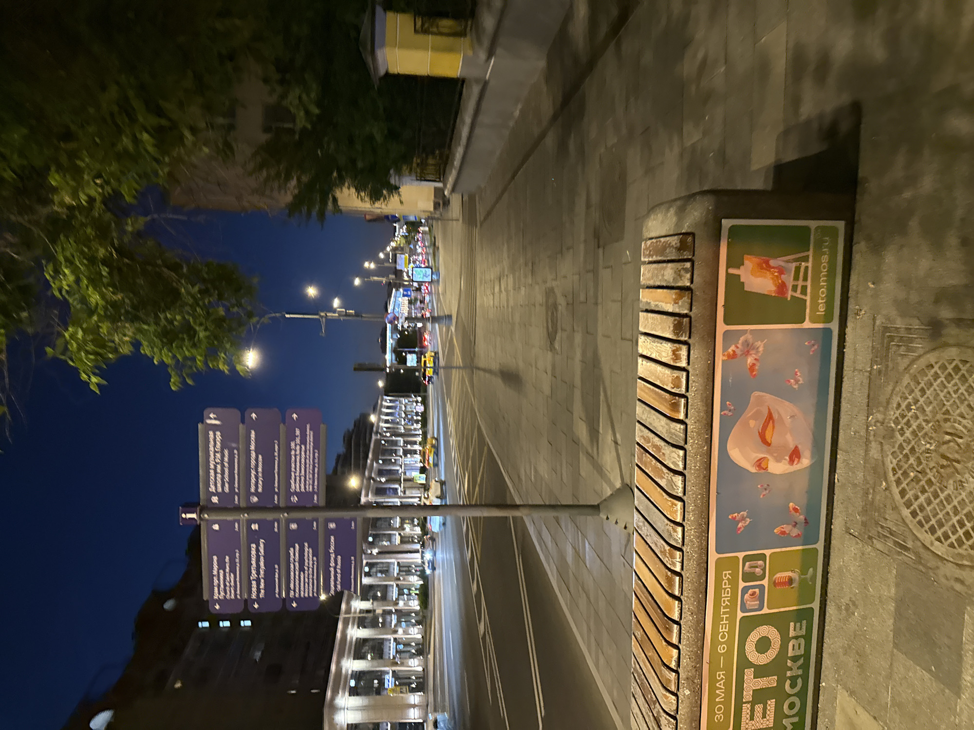 This image captures a wide-angle view of a city street in Moscow, Russia, at night or late evening. The specific location is **Bolshaya Polyanka Street**, indicated by the prominent building on the left side of the street which closely resembles the "Dom na Polyanke" complex, as well as the destinations listed on the signpost.
The scene depicts a multi-lane asphalt road illuminated by numerous streetlights, separating the viewer from a large, modern building complex. This building features multiple stories with illuminated arched windows and columned facades. Several vehicles, including cars and yellow taxis, are visible on the road, some with headlights on, suggesting traffic flow.
In the foreground on the right, a paved sidewalk stretches into the distance. A sturdy concrete bench with wooden slats on top occupies the lower part of the frame. The side of the bench displays a colorful advertisement for "ЛЕТО В МОСКВЕ" (Summer in Moscow), dated "30 МАЯ – 6 СЕНТЯБРЯ" (May 30 – September 6), featuring icons for photography, music, a microphone, an artistic mask with butterflies, and an easel, along with the website "leto.mos.ru".
Just beyond the bench, a tall, grey metal signpost stands prominently, bearing multiple purple directional signs with white text in both Russian and English. These signs point to various local landmarks and institutions:
* "Храм прп. Марфо-Мариинская / Church of Saint Martha, the Merciful" (3 min away)
* "Новая Третьяковка / The New Tretyakov Gallery" (2 min away)
* "Социальный фонд России / Social Fund of Russia"
* "Детская музыкальная школа им. В.М. Глиэра / Glier School of Music"
* "Нотариус города Москвы / Notary in Moscow"
* "Судебные участки № 386, 387 / Judicial Sections No. 386, 387"
Further along the sidewalk to the right, a lower, yellow-colored building with a dark fence and a grey stone wall is partially visible, obscured by green trees. The sky above is dark blue, characteristic of night, with no clouds. The overall lighting is provided by streetlights, vehicle headlights, and the illuminated building facades. No people are clearly visible in the frame. A metal manhole cover is visible on the sidewalk in the lower right corner.