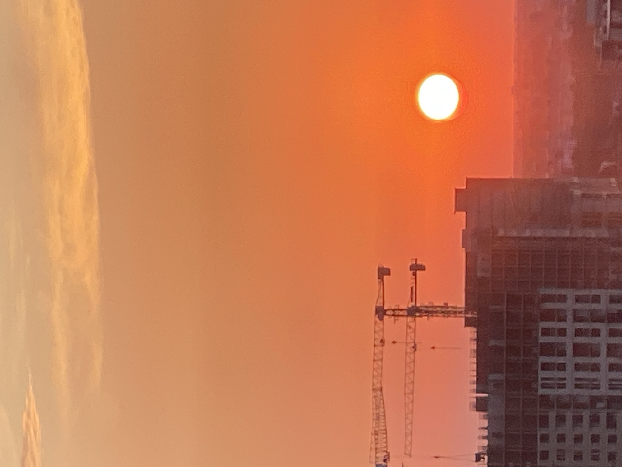 An aerial, vertical, full shot captures a sunset over an urban skyline. The sky is a vibrant orange, with lighter orange clouds near the top. A large, bright white sun is visible in the upper right quadrant of the image. In the mid-ground, the dark silhouettes of several construction cranes are visible, with one crane in focus in the center. The cranes are positioned above a partially constructed, large building. This building is composed of concrete and steel, with visible scaffolding and unfinished floors. The building occupies the lower left and center of the image. The background shows a hazy, distant city skyline. The overall impression is one of ongoing urban development against a dramatic sunset.