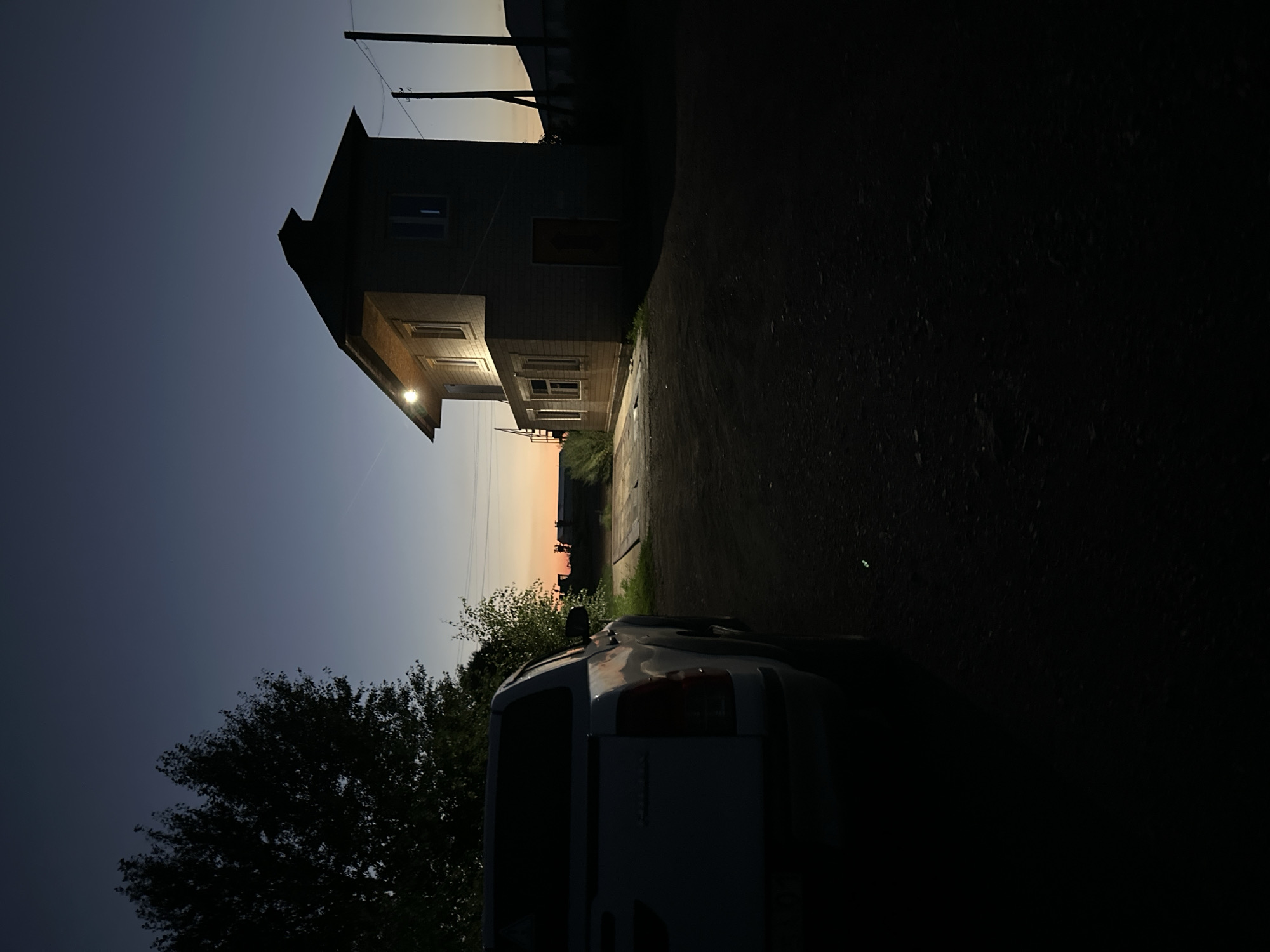 A white pickup truck is parked on a dirt road facing away from the camera on the left side of the frame. To the right, a two-story building with a pitched roof is illuminated by a single light fixture on the upper facade. The building appears to be constructed of light-colored brick or siding. A metal staircase is visible on the left side of the building. In the background, a dark silhouette of trees is present on the left, and a bright orange and yellow sunset is visible on the horizon. Telephone poles and wires are also visible in the distance. The scene is set at dusk, with the sky transitioning from a deep blue to a lighter hue near the horizon.