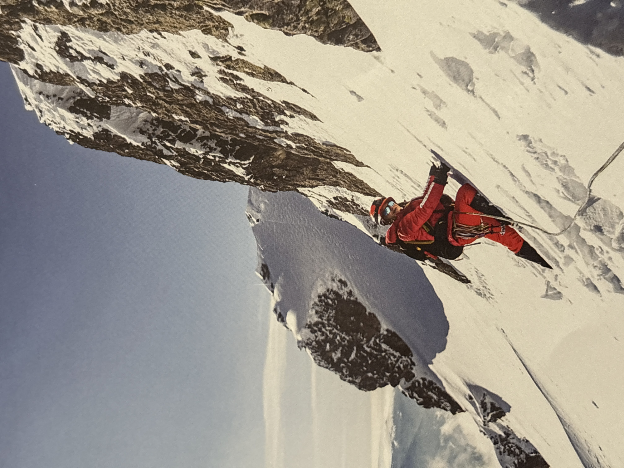 A climber in red gear, wearing a helmet and goggles, is ice climbing on a steep, snow-covered mountain face. A rope extends upwards from the climber. The sky is visible in the lower left corner. The location is not within the city of Plovdiv, Bulgaria.