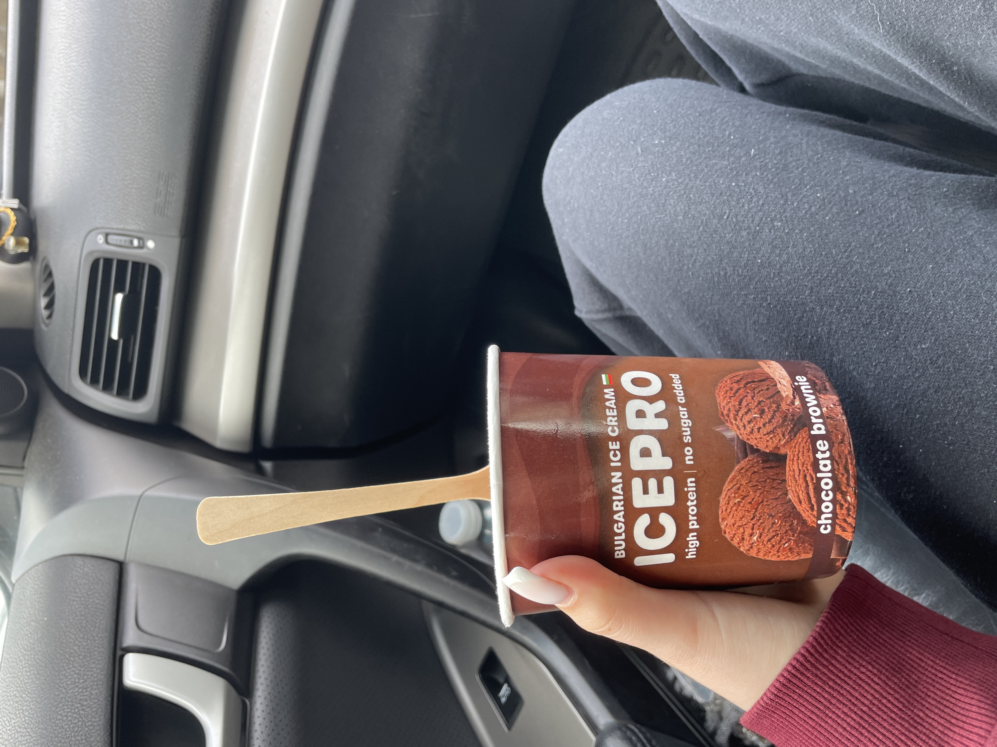 A person inside a car is holding a container of "ICEPRO" Bulgarian ice cream in the flavor "chocolate brownie." The container advertises "high protein" and "no sugar added." A wooden spoon is sticking out of the container. The person is wearing a long-sleeved red shirt. The interior of the car is visible, including the dashboard and door panel. The location in the city of Plovdiv, Bulgaria, is not explicitly stated in the image.