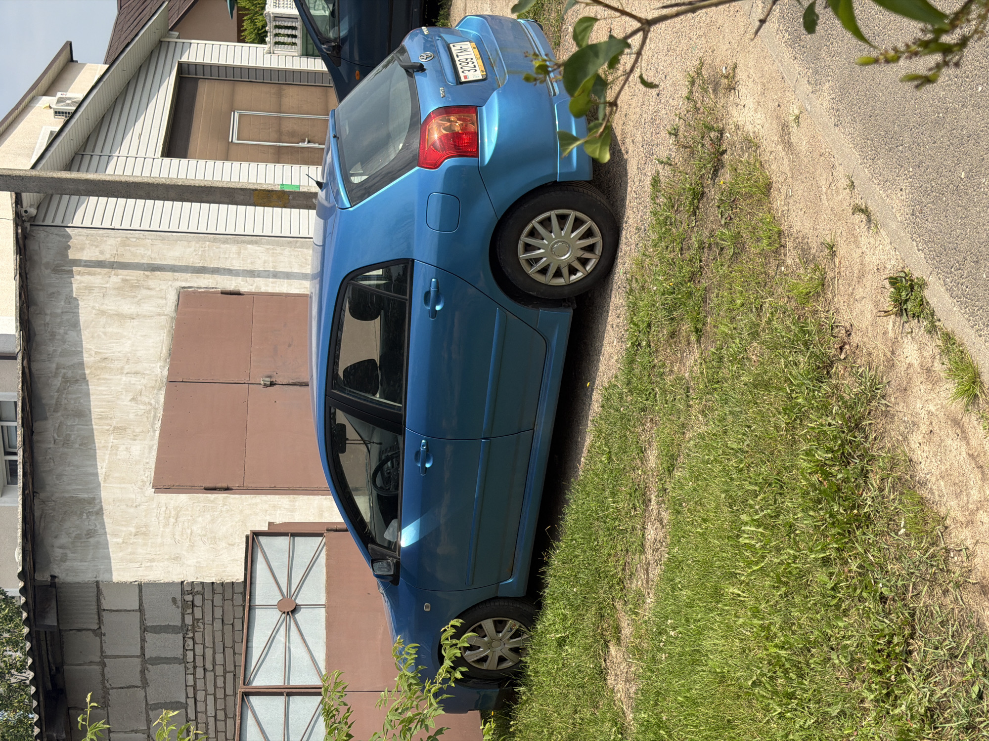 The image shows a blue Toyota hatchback parked on a gravel and grass area in front of a building. The building appears to be a garage or workshop with a large, brown metal door. To the right of the main building is another section with white siding and a smaller brown garage door. The car's license plate is visible, reading "3299 TM-7". The scene is outdoors on a sunny day, with green grass and some trees. Based on the license plate format and the overall aesthetic, the location is likely Minsk, Belarus. There are no people visible in the image.