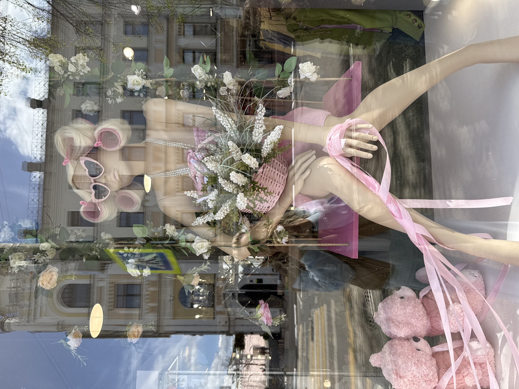 The image shows a window display in Заволжье, Russia. The display includes a mannequin with blonde hair in curlers, pink heart-shaped sunglasses, and a pink outfit. The mannequin is holding a pink basket filled with white flowers and greenery. Pink ribbons are draped around the mannequin's arm and are accompanied by pink teddy bears. The window's reflection shows a building and a street sign. The overall aesthetic of the display is pink and floral.