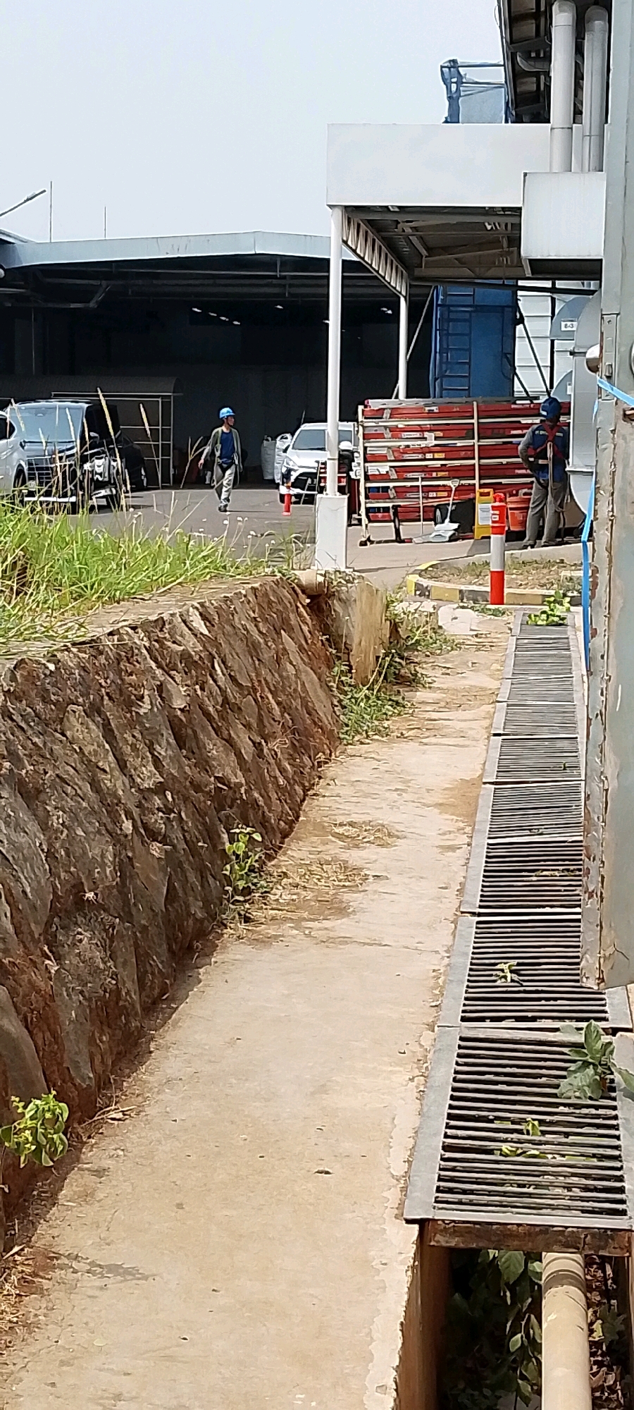 This image depicts an outdoor scene within an industrial or construction area, possibly located in Depok, Indonesia, given the provided context. In the mid-ground, a person wearing a blue hard hat and a light-colored jacket over a dark shirt and pants is walking on a paved path. To the right, another person in similar work attire, including a hard hat, is standing near a large stack of red planks or materials, which are arranged horizontally. Further to the left, two cars are parked, one with its hood open, and a dark structure resembling a warehouse or large building with a sloped roof is visible in the background. A concrete wall with a rough, rocky texture lines the left side of the path. On the right, a grated drain runs along the edge of the path, with some vegetation growing around and within it. The overall environment appears to be functional and under development or in operation, with elements of construction and infrastructure present.