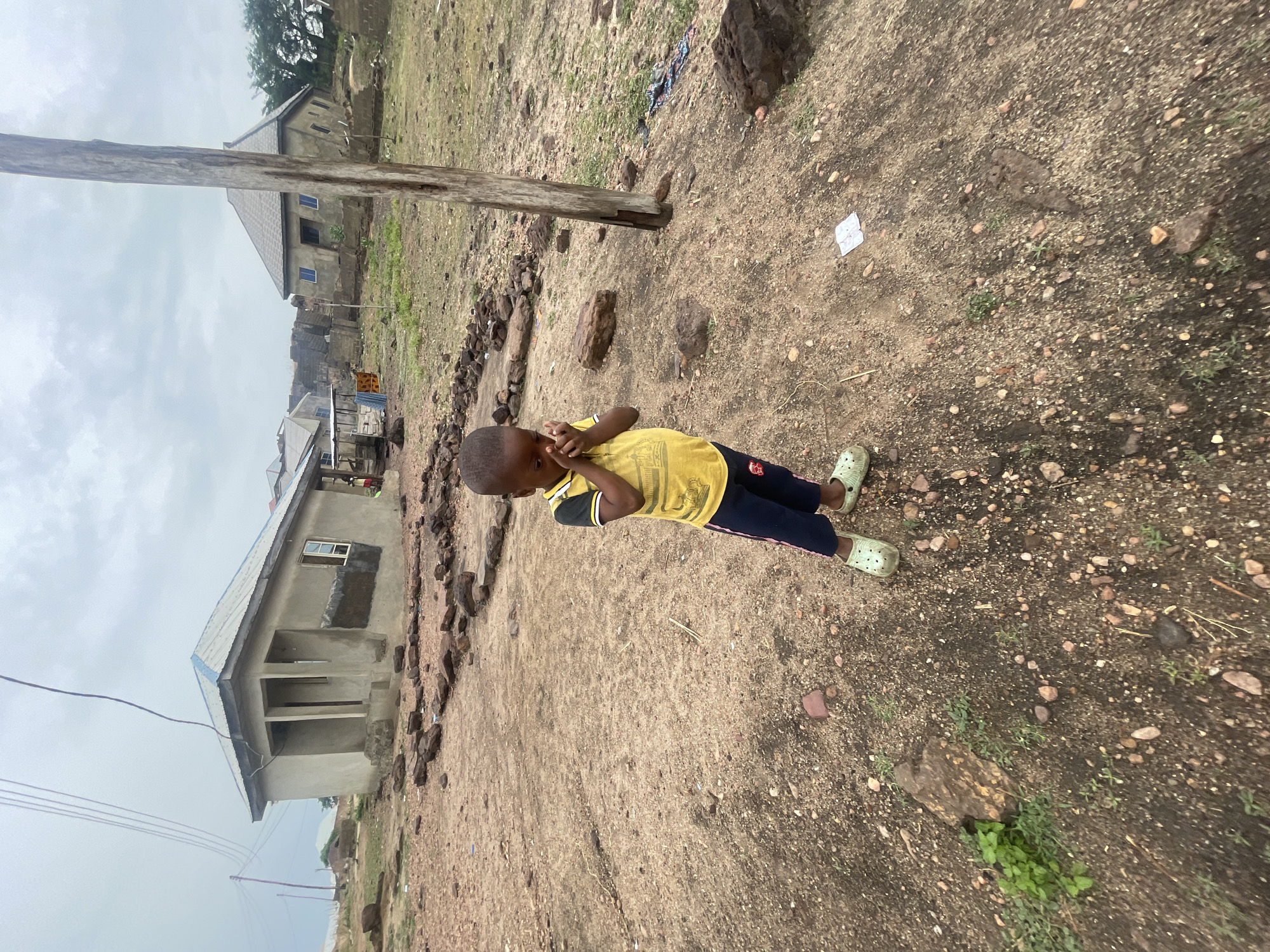 A full-body shot shows a child standing outdoors in a somewhat barren area. The child, wearing a yellow t-shirt and dark pants, has their hands near their face. A wooden pole stands near the child. In the background, there are several buildings, including what appears to be a house under construction. The ground is dirt and gravel, with some sparse vegetation. The photograph suggests an overcast day. The image is consistent with a rural setting in a developing area. It's difficult to definitively pinpoint this area as Lokoja, Nigeria, without further evidence, but it fits the general description of some parts of Lokoja.