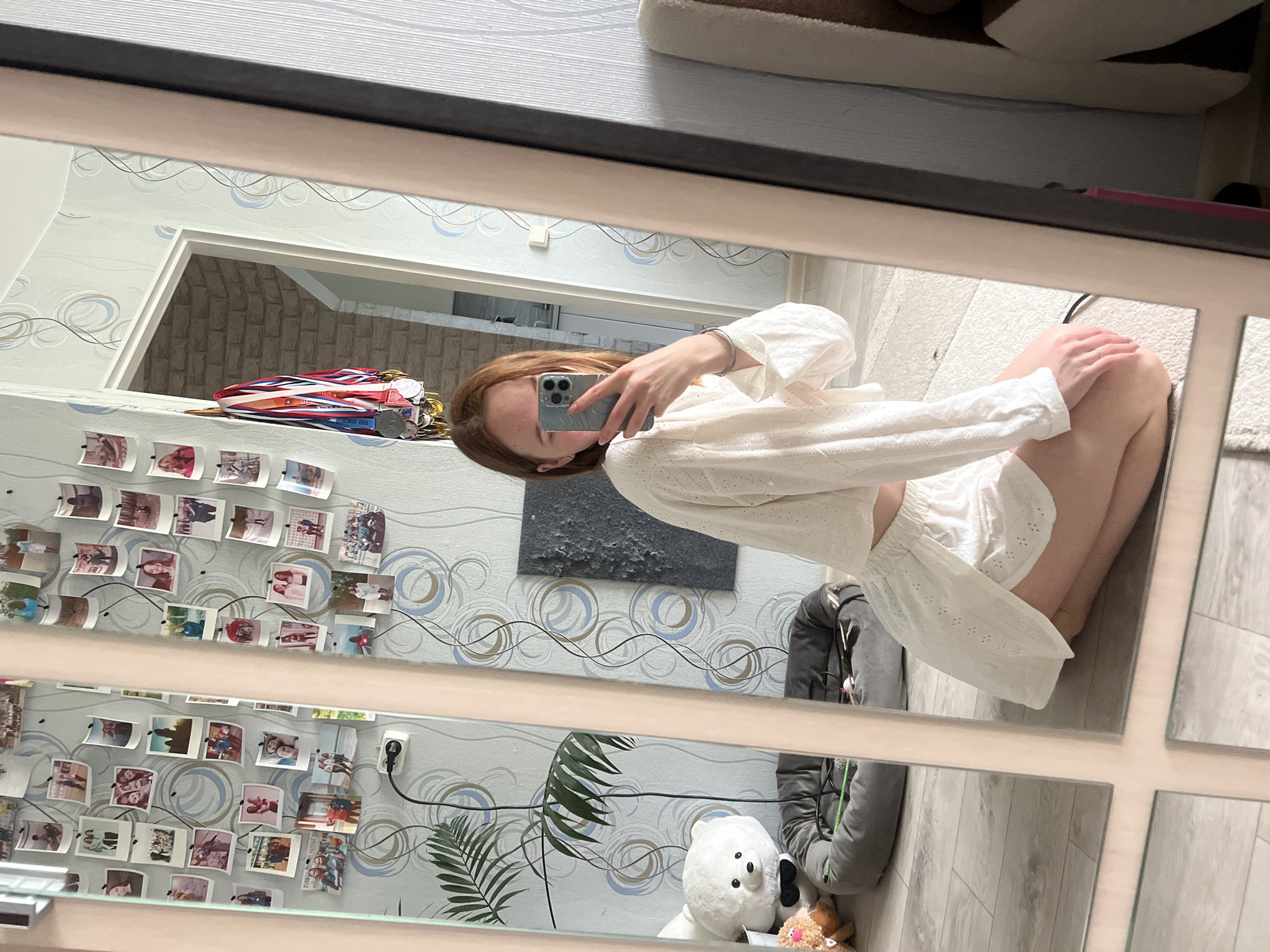 A young woman is taking a selfie in a mirror, the reflection of which shows her room. The room is decorated with wallpaper featuring a pattern of circles and lines, with photos pinned to the wall. A teddy bear and a gray jacket are visible on the floor. Medals hang near a doorway. The location is Chaikovsky, Russia.