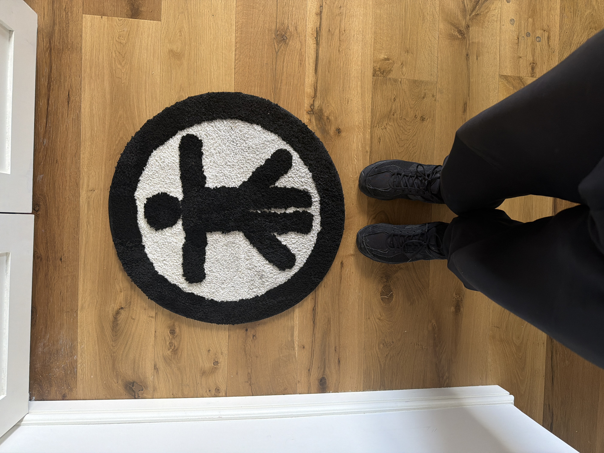 The image shows a section of a room with light brown wooden flooring. In the center-left of the frame lies a circular rug with a black border. The rug features a white background with a black figure resembling a stylized human form within the circle. To the right of the rug, a pair of black athletic shoes and black pants are visible, suggesting someone is standing near the rug. A partial view of a white door frame is seen on the left side of the image. The lighting appears to be natural and even. No information in the picture suggests a location in the city of London, United Kingdom.
