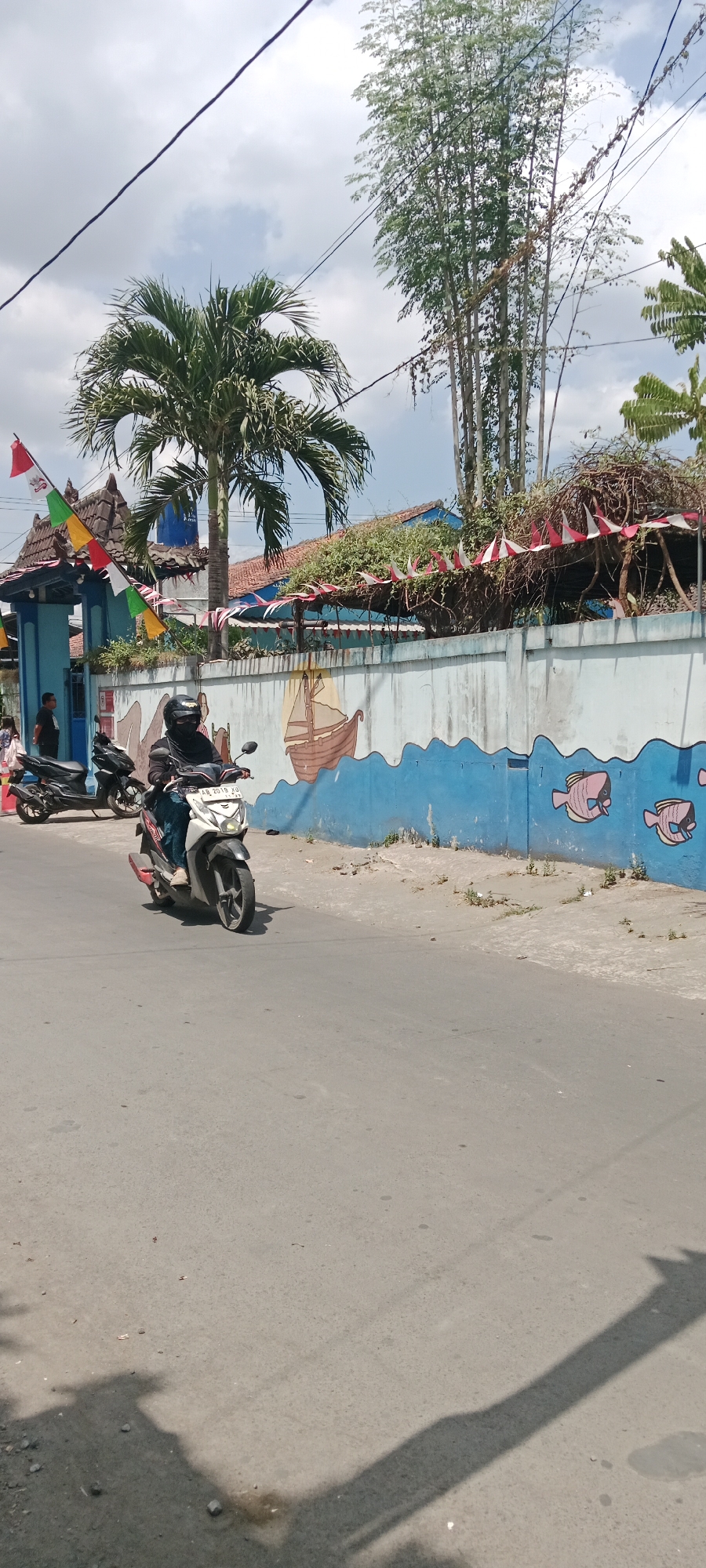 In Purwomartani, Indonesia, a person wearing a black hijab and helmet is riding a white and black scooter on a paved road. The scooter has a license plate reading "AB 2018 XU". In the background, a blue wall is decorated with a mural depicting a ship and fish. Red and white festive flags are strung along the top of the wall and above an entrance gate. A palm tree stands near the gate. To the left of the scooter, another black scooter is parked. Two people, one wearing a black shirt and another in a pink shirt, are standing near the entrance. The sky is partly cloudy.