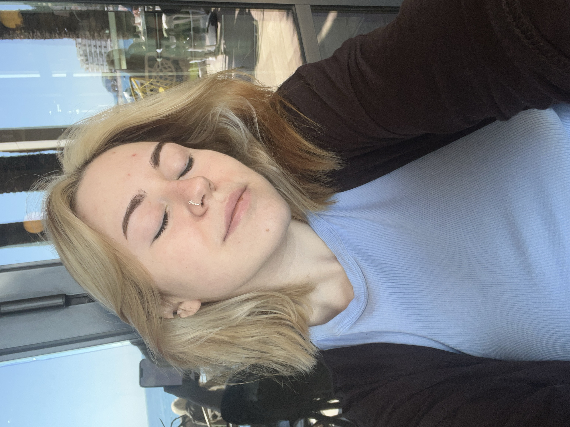 A young woman with blonde hair and a nose ring is lying down, eyes closed, wearing a blue ribbed shirt and a dark brown jacket. The background shows a reflection of buildings and a bright sky, possibly seen through a window. The reflection suggests an urban setting near the coast, possibly in a coastal area with high-rise buildings. There is no evidence to place her specifically in Rafailovići, Montenegro, based on this image.