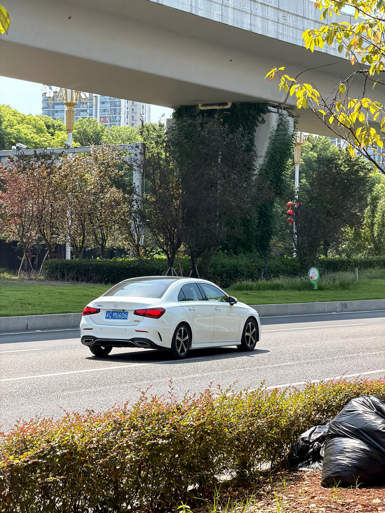A white Mercedes-Benz A-Class sedan with license plate "C MU606" is driving on a paved road under an elevated concrete structure. The car is positioned in the right lane of a two-lane road. The road is bordered by a grassy median strip and a dense hedge in the foreground. To the left of the road, a manicured lawn is visible, with trees and buildings in the background. The sky is clear and blue. The scene appears to be a street in the city of Jiangjin, China. Two black garbage bags are present on the ground to the right of the hedge.