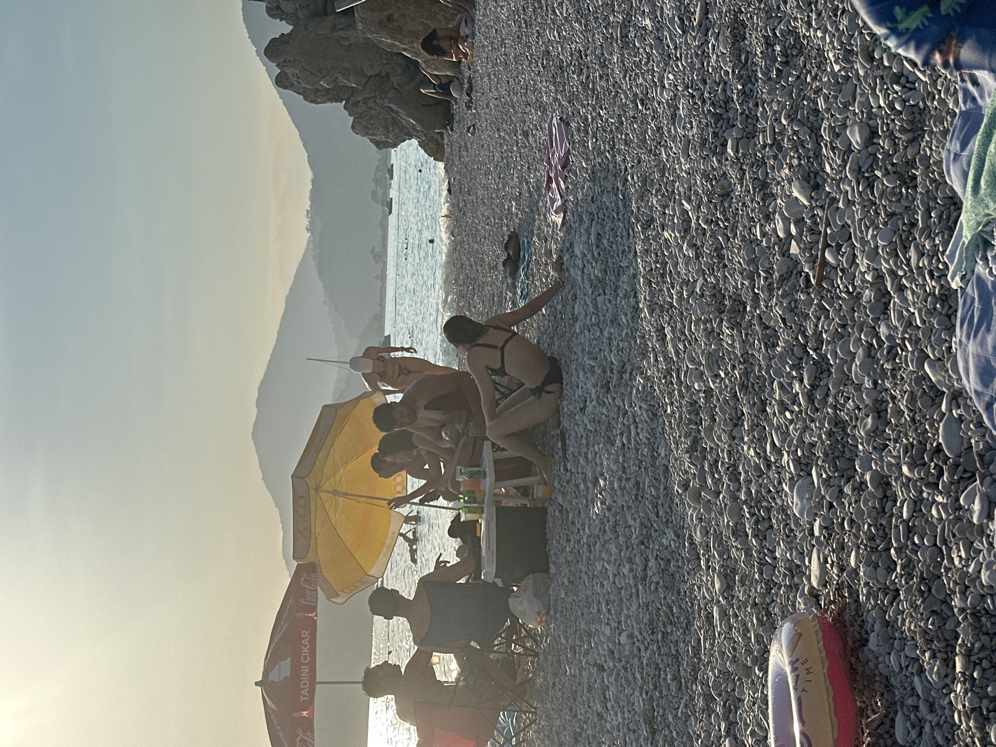 This image captures a late afternoon or sunset scene on a pebble beach in the Fethiye region of Turkey. The environment features a wide expanse of light grey and white pebbles forming the beach, extending into the foreground. The calm, blue-grey sea sparkles with sunlight reflections towards the horizon. In the background, multiple layers of hazy, silhouetted mountains rise, partially obscuring the distant sky. A rocky outcrop is visible on the far right side of the beach. The sky transitions from a bright, hazy yellow-white near the horizon to a lighter blue overhead, indicative of low sun angle and atmospheric conditions.
Several individuals and groups are present. In the mid-ground left, a group of approximately five to six people is gathered under two beach umbrellas: a red Coca-Cola branded umbrella displaying the Turkish text "TADINI ÇIKAR" (Enjoy the taste), and a yellow umbrella. This group is seated on portable chairs or directly on the pebbles around a small, low white table. The table holds several drinks in bottles, cans, and a glass containing an orange liquid. A dark-colored cooler and a white plastic bag are positioned near the table. The individuals in this group are engaged in conversation and relaxation; most are partially silhouetted against the bright water.
Further right in the mid-ground, a woman in a dark bikini top and bottom is seated on the pebbles, facing away from the viewer, leaning on her left arm while looking towards the sea. To her left, partially obscured, a person wearing a light-colored cap and a patterned bikini is walking out of the water onto the beach. Several other indistinct figures are visible swimming or wading in the sea, and a few small boats are anchored further out in the bay. Near the rocky outcrop on the right, additional individuals are visible sitting on towels or blankets on the beach.
In the immediate foreground, a pink and yellow inflatable swim ring with the word "TIME" visible is resting on the pebbles to the left. A blue patterned blanket or towel is partially visible in the bottom right corner, suggesting another person is situated just out of frame. The overall atmosphere is one of leisure and relaxation by the sea during the golden hour.