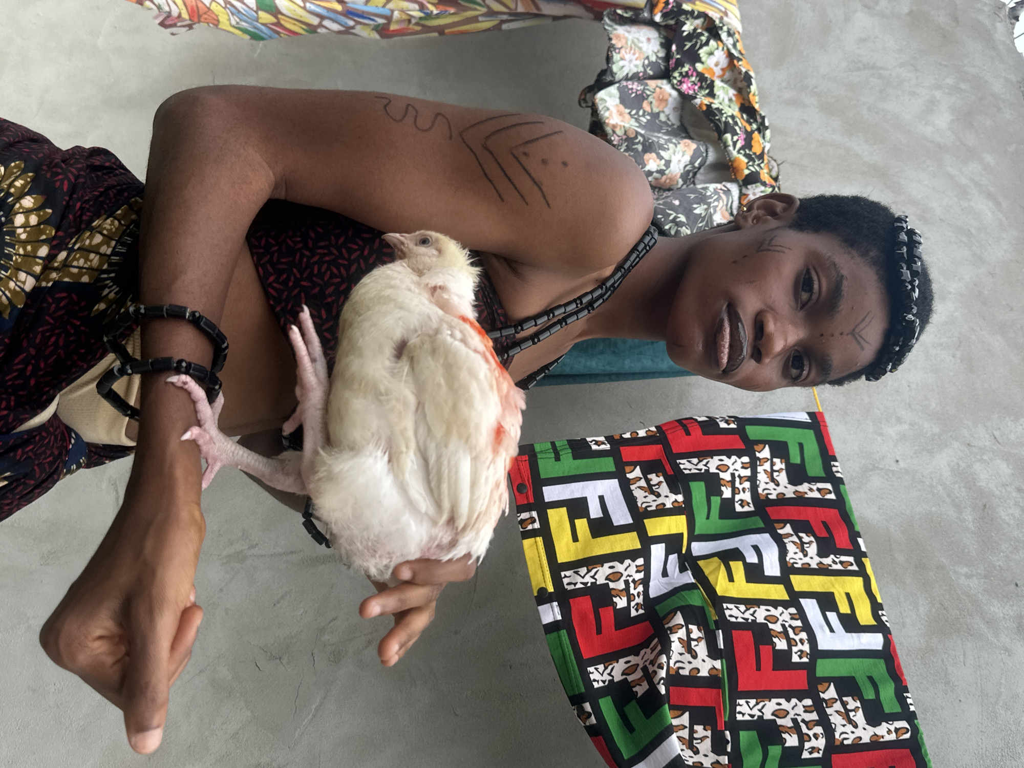 In an outdoor setting against a grey concrete wall in Badagry, Nigeria, a young woman with dark skin and short hair, adorned with intricate face markings and a beaded headband, holds a white chicken. She is wearing a black and red patterned top and a dark, patterned skirt. Her left arm, which is tattooed with geometric designs and a wavy line, supports the chicken. A vibrant fabric with the "FF" logo and leopard print is draped behind her to the right, and to her left, another patterned cloth hangs. Her posture and the way she holds the chicken suggest a deliberate pose for the photograph.