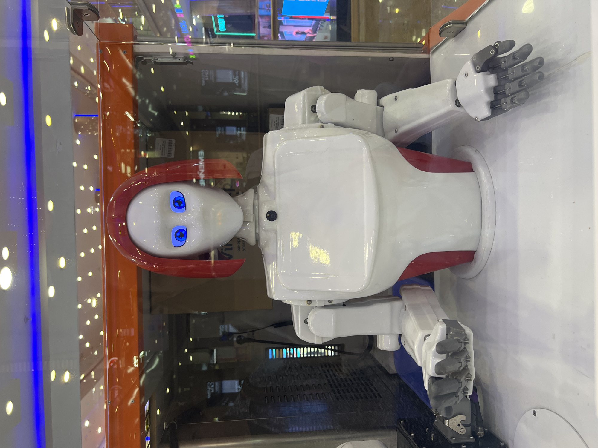 This is a medium shot, eye-level photograph of a humanoid robot. The robot is white with red accents and has bright blue eyes. It is positioned behind a glass enclosure in what appears to be a shopping mall or electronics store. The background is blurred but shows displays with lights and reflections, indicating an indoor commercial environment. The robot's arms are extended, and its hands are open, with one hand showing a more complex articulated structure with visible finger joints and metallic accents, while the other hand appears to be a simpler claw-like gripper. The overall impression is of a modern, potentially interactive exhibit.