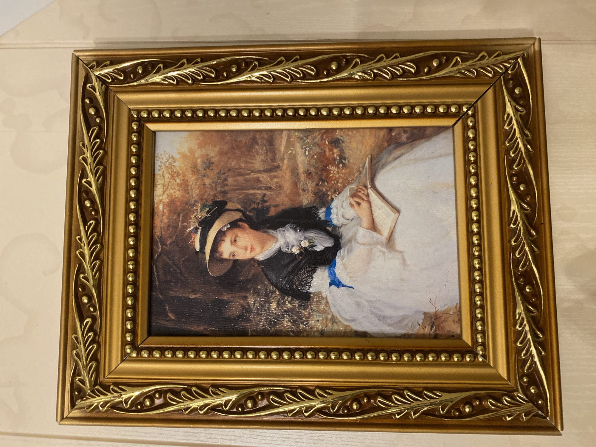 A framed rectangular portrait of a young woman is displayed centrally on a light-colored, subtly textured wall. The painting, rendered in a realistic, possibly Impressionistic style, depicts a Caucasian woman seated outdoors in an autumnal wooded or park setting. She wears a wide-brimmed straw hat with a dark band and decorative elements, a white dress with a ruffled high neckline, a dark shawl or short jacket over her shoulders, and a prominent blue ribbon detail on her dress. Small white flowers, possibly daisies, are pinned to her bodice. Her hands are clasped over an open book resting on her lap, and she gazes with a contemplative expression slightly upwards and to the viewer's left. The background of the painting features trees with foliage in shades of brown, orange, and yellow, with the ground covered in dry grass and scattered vegetation.
The artwork is enclosed within an ornate, gold-colored frame. The frame features a decorative raised relief pattern of intertwined leaves and small beads along its outer perimeter, and a continuous row of smaller, spherical raised beads along its inner perimeter, directly bordering the painting. The corners of the frame are mitered at a 45-degree angle. The wall behind the frame is a uniform light hue, possibly off-white or cream, and shows faint vertical lines and a subtle, embossed-like pattern, suggesting wallpaper or textured paint. No specific city or geographic location can be determined from the image.