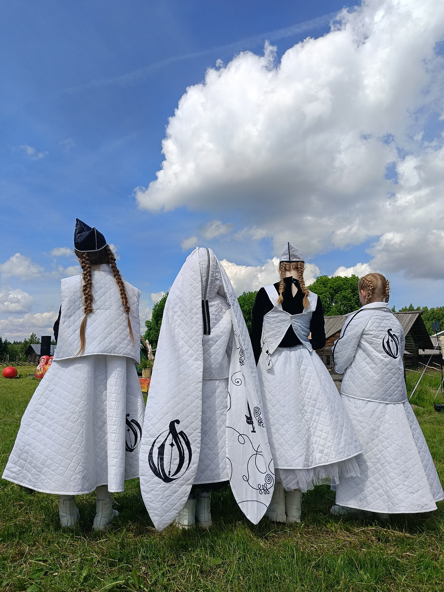 Four individuals, viewed from behind, stand on a grassy field under a partly cloudy blue sky. They are dressed in matching quilted white outfits consisting of capes, skirts, and hats. The outfits are adorned with black emblems, one of which resembles a stylized onion or droplet. The individual on the far left wears a dark blue cap with a white trim. The individual second from the left is almost entirely obscured by a large, quilted white cape that hangs to the ground, revealing only white boots. The individual second from the right wears a black top with white trim over a white skirt and a grey and white patterned cap. The individual on the far right wears a quilted white jacket and skirt, with a black emblem on the back of the jacket, and a grey and white patterned cap. In the background, trees, wooden structures, and a red sphere are visible. The scene appears to be outdoors, possibly at a festival or event in the village of Popovka, Russia.