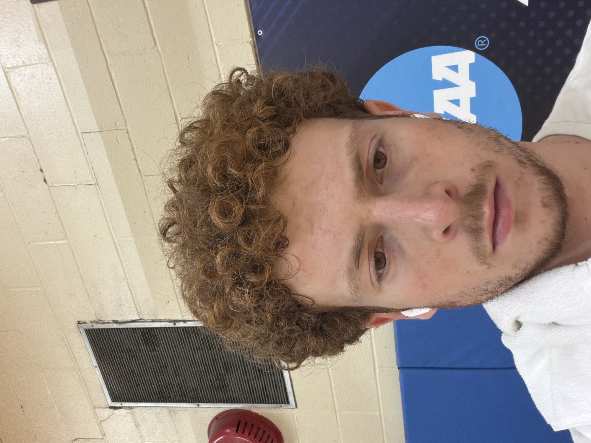 A close-up, portrait-style shot shows a man indoors. He has light brown, curly hair and light brown eyes, with light stubble on his face. White earbuds are visible in his ears, and a white towel is draped over his right shoulder. He looks directly into the camera with a neutral expression.
The background features an off-white cinder block wall on the left side and a dark blue banner or panel on the right. A prominent blue circular logo with white text, clearly displaying "NCAA" and a registered trademark symbol (®), is visible on the dark blue background. Part of "AA" within the logo is also distinguishable. Below the banner, another section of a blue mat or panel is present. On the lower left, a rectangular ventilation grate is mounted on the wall, and beneath it, a portion of a red, possibly bell-shaped object is visible. The setting suggests an indoor sports or educational facility, likely a gym or common area. This scene is situated in New York, United States of America.