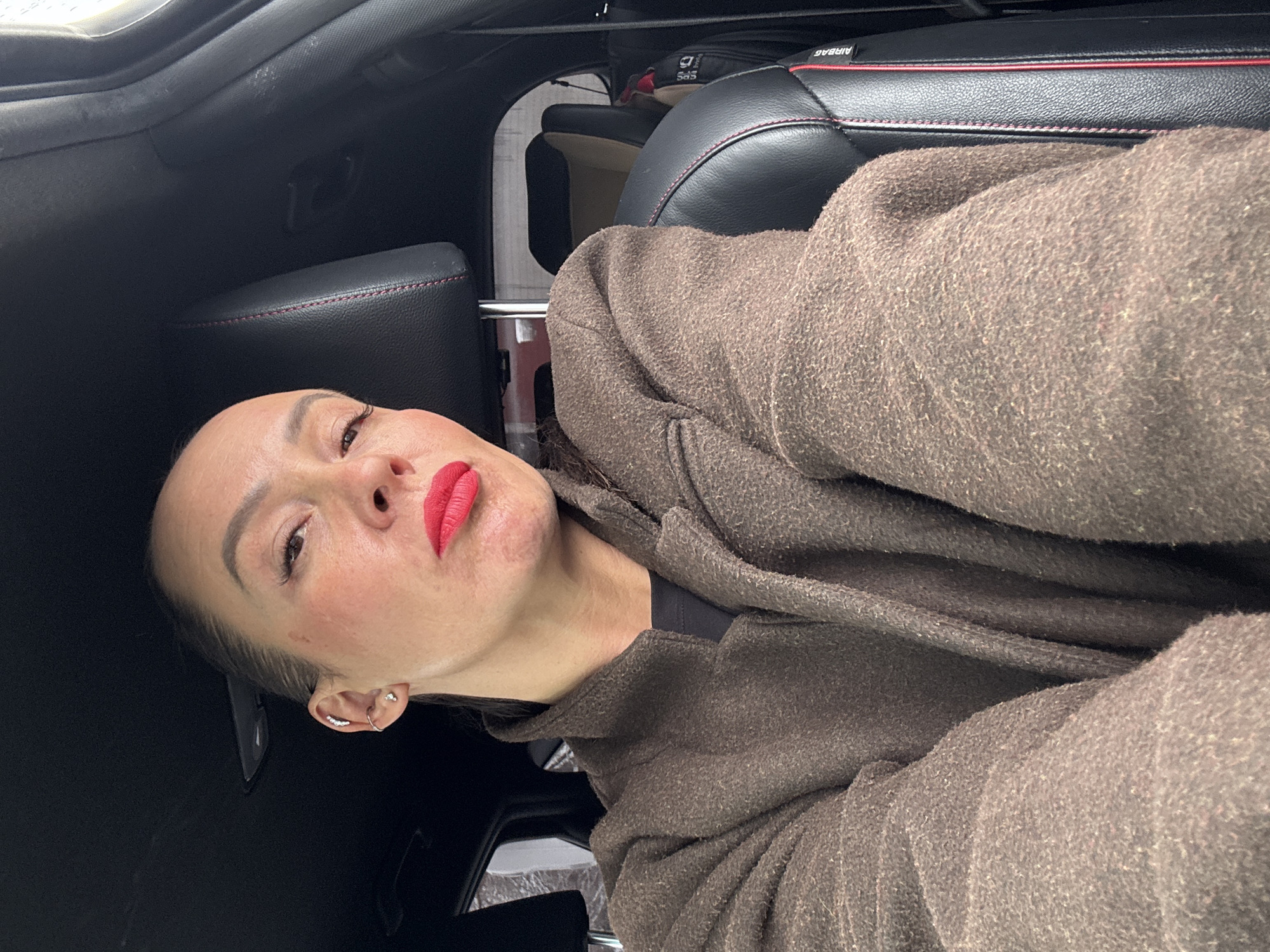 A woman is inside a car. She wears a brown coat and bright red lipstick. The car has black leather seats with red stitching. The location is not discernible from the image.