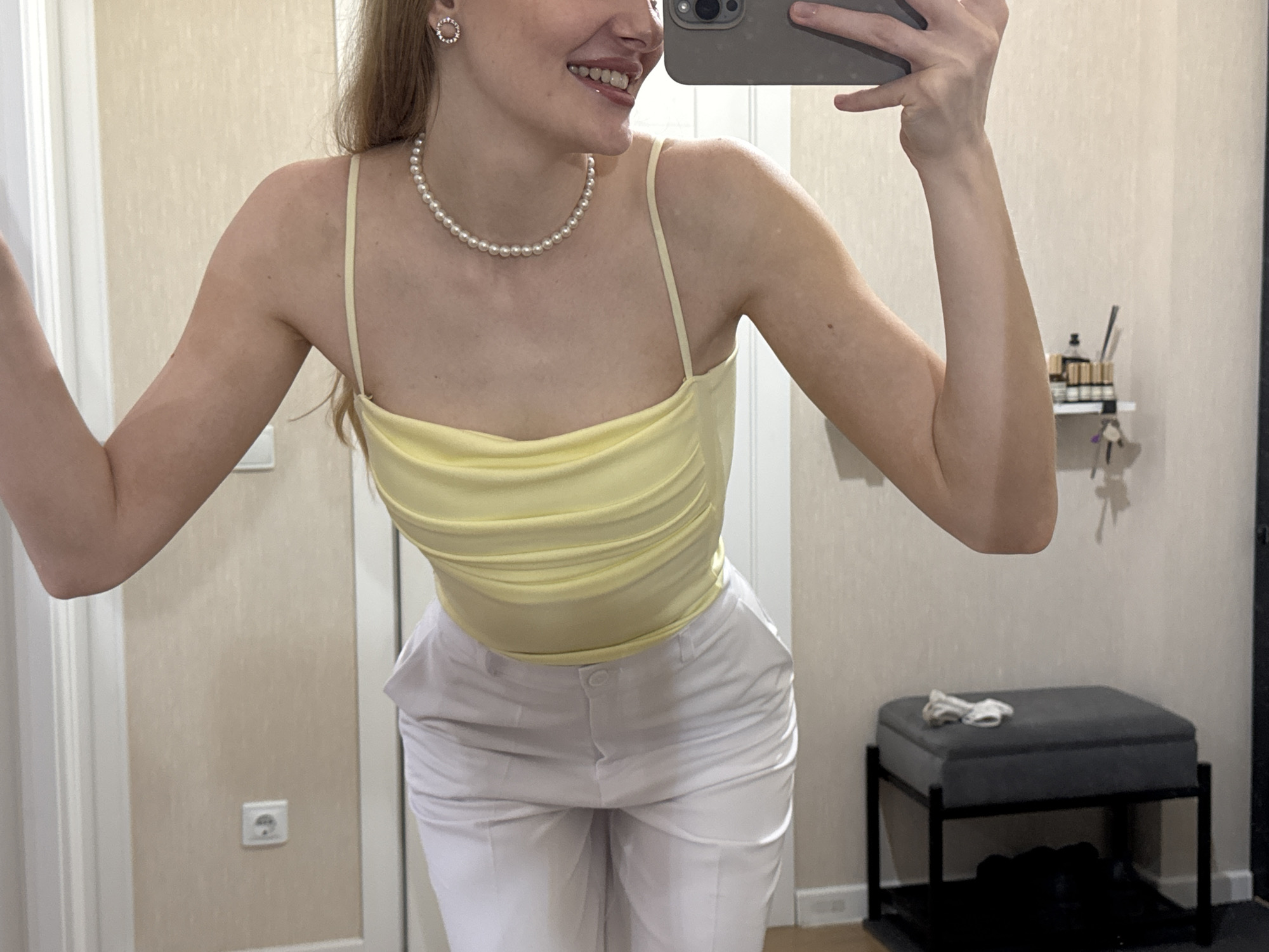 The media file shows a person taking a mirror selfie in a room. The person is wearing a light yellow ruched camisole and white pants. They have on a pearl necklace and circular earrings. The background suggests an indoor setting, possibly a bedroom or dressing area, with a textured wall, a doorframe, and an electrical outlet visible. To the right, there is a shelf with bottles and a grey ottoman with some fabric on top. The lighting appears to be artificial, consistent with an indoor environment. The person is smiling, indicating a positive or playful mood. The presence of a mirror and the way the person is holding a phone suggests the activity is taking a picture of themselves. The location context provided is Perm, Russia, but this is not directly evident from the visual information in the image.