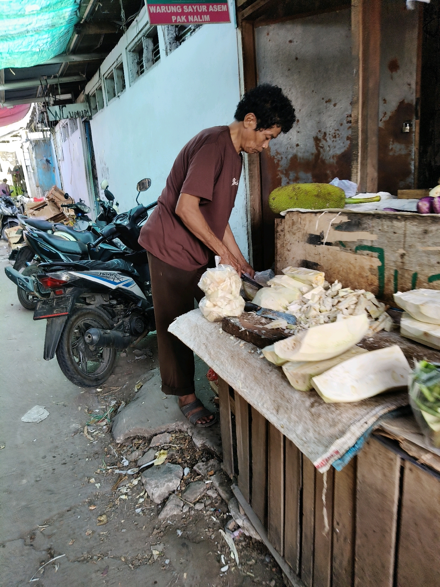 In Setiamekar, Indonesia, a person is observed at a market stall, chopping what appears to be unripe jackfruit. The individual, wearing a brown t-shirt and brown pants, is focused on the task at hand, with a knife in their hands and a pile of chopped jackfruit in front of them. A large, whole unripe jackfruit sits on the counter to the right of the person. The stall itself is made of wood and displays various cut pieces of the jackfruit, some still intact. In the background, several motorcycles are parked, suggesting a busy area. The ground around the stall is uneven and shows signs of wear, with scattered debris. A sign above the stall reads "WARUNG SAYUR ASEM PAK NALIM," indicating the name of the food stall and likely the proprietor.