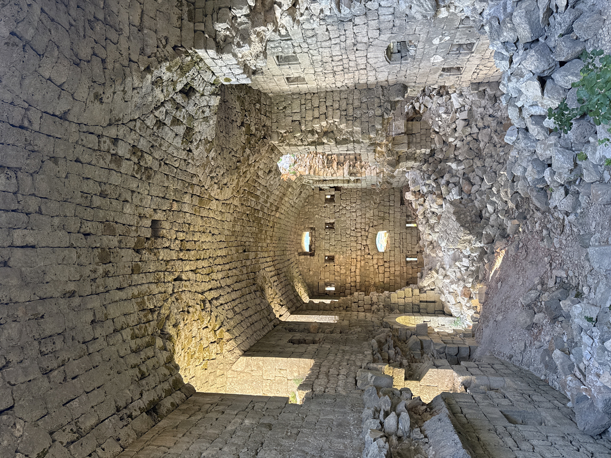 The image shows the interior of a stone structure, likely a ruin, with a high, arched ceiling constructed from meticulously laid stones. Sunlight streams in from openings, illuminating piles of rubble and debris that have accumulated on the floor. The walls are also made of stone, showing signs of damage and decay, with some sections crumbled away. Small, rectangular openings are visible in the walls, suggesting former windows or embrasures. The overall impression is one of ancient construction and the passage of time, with nature beginning to reclaim the man-made structure. The location is identified as being in the city of Brajici, Montenegro.