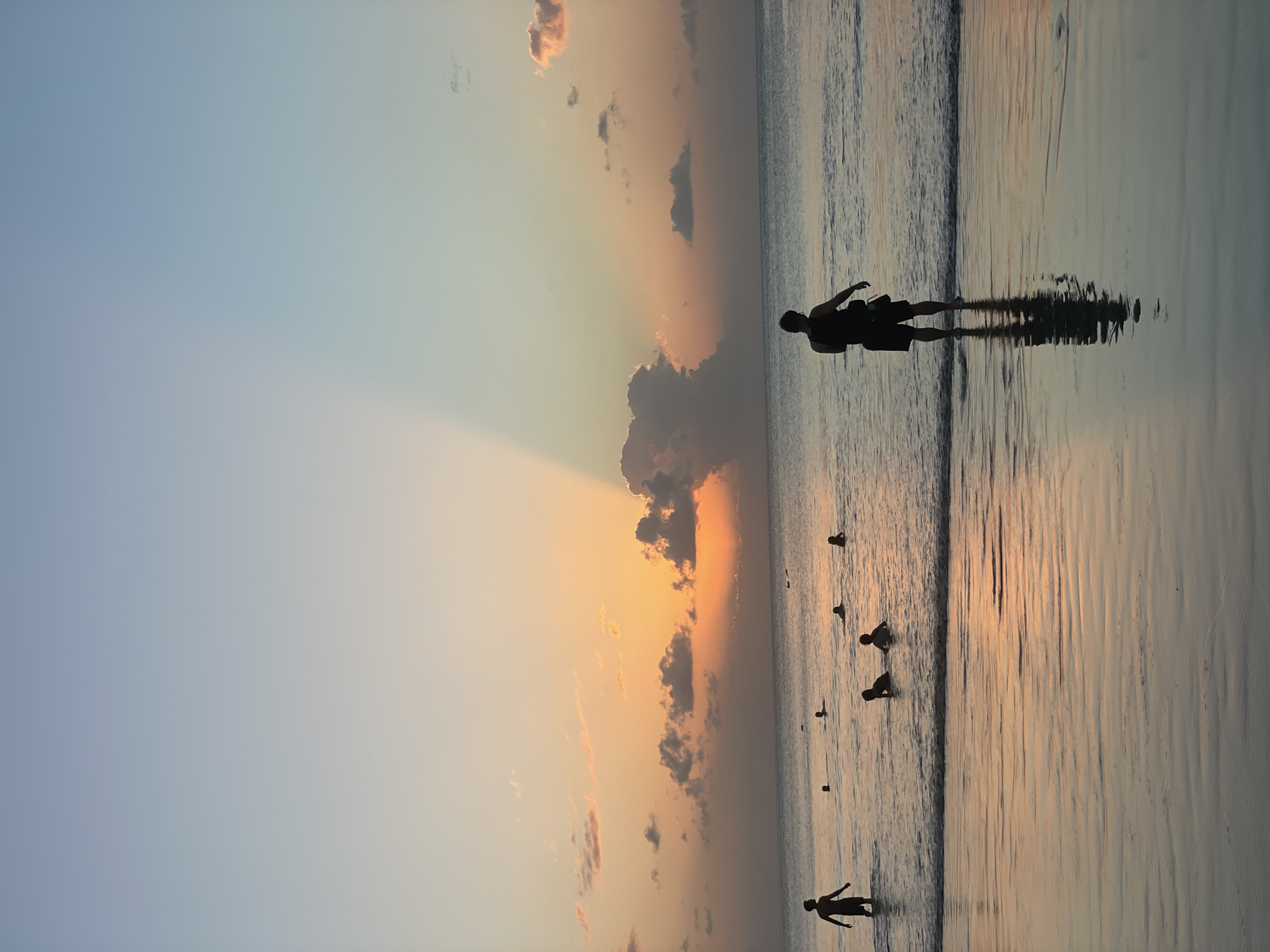 A photograph captures a sunset over the ocean with several people wading and swimming in the shallow water near a sandy beach. The sky displays a gradient of soft blues and oranges, with scattered clouds illuminated by the setting sun. A distinctive sunbeam effect splits the sky. In the foreground, the wet sand reflects the colors of the sky. Two prominent silhouetted figures are visible in the water: one on the left, facing the ocean, and another on the right, facing away from the camera. Several other indistinct figures are scattered throughout the water. The scene is likely Kuta Selatan, Indonesia, known for its popular beaches and sunsets.