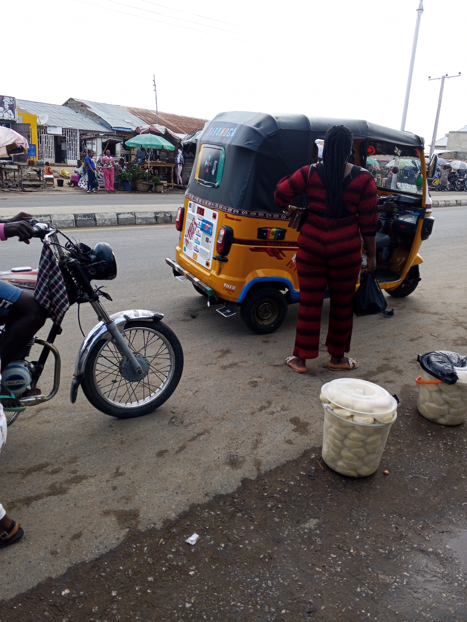 This image captures a busy street scene in Zango-ndaji, Nigeria, under an overcast sky. The setting features a paved road with a raised curb, alongside which are several market stalls and shops with corrugated metal roofs.
In the foreground on the left, a person, partially visible, sits on a black and silver Bajaj motorcycle. A plaid cloth is draped over the motorcycle's handlebars. In the mid-ground, a woman with long black braids, dressed in a red and black striped jumpsuit and flip-flops, stands with her back to the viewer. She holds a black plastic bag and appears to be interacting with or waiting by a yellow "keke napep" (auto-rickshaw) with a black canopy. The tricycle has the word "DELUXE" on its side, and some text that looks like "ZORU..." and "TOJU..." is visible on the rear, along with various stickers.
To the right of the woman, two clear plastic buckets with white lids are placed on the ground, filled with what appear to be peeled food items, possibly yams or similar produce, suggesting street vending. The road surface shows patches of moisture and some debris.
In the background, behind the tricycle, numerous people are gathered around market stalls, some covered by green and white umbrellas. A sign on one of the stalls faintly reads "THANKS FOR YOUR PATRONAGE." Utility poles and wires are visible against the light, cloudy sky. The overall scene depicts a typical bustling commercial street or market activity.