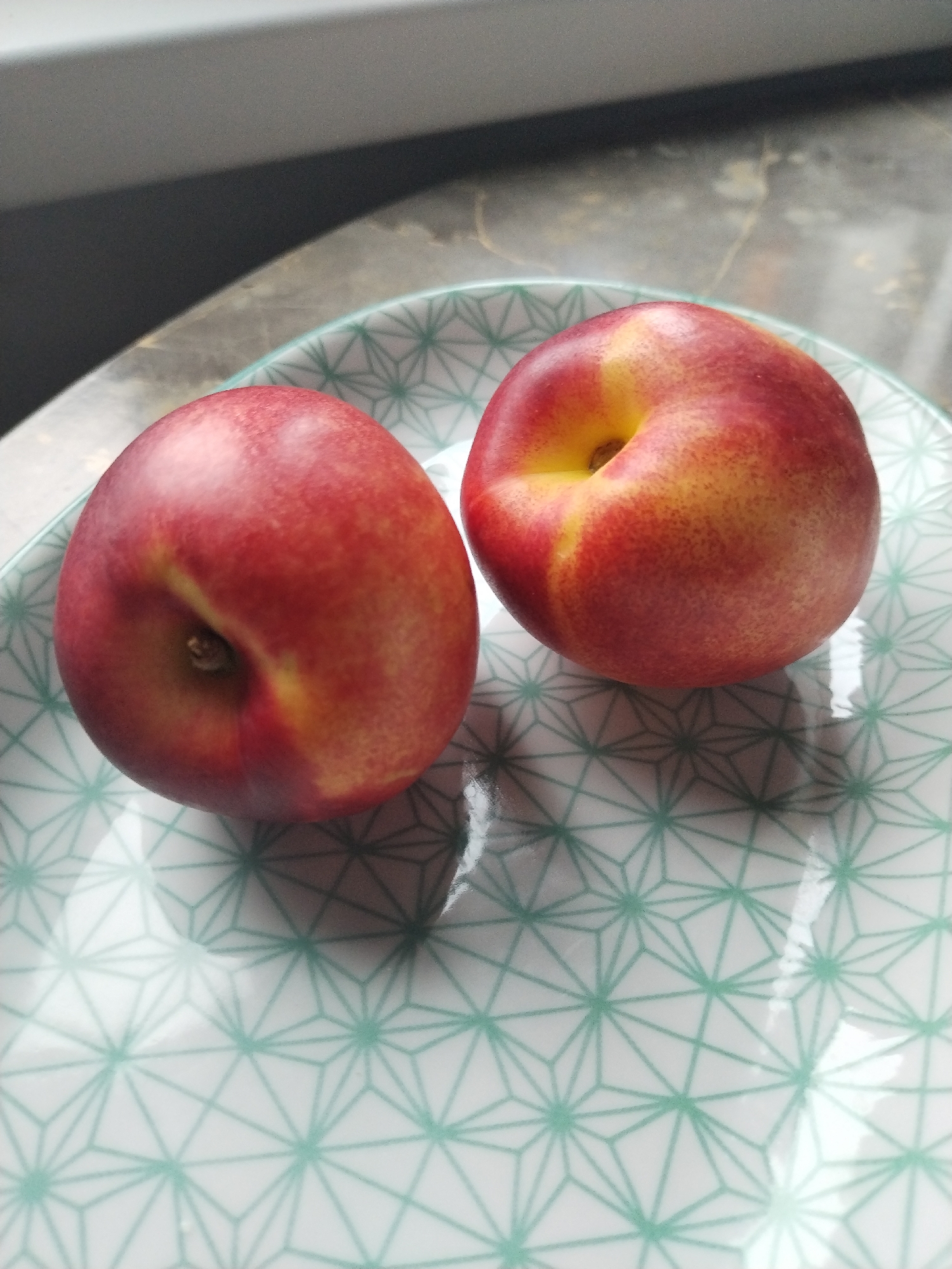 Two whole nectarines are centrally positioned on a white ceramic plate adorned with a teal-green geometric pattern. Each nectarine displays a smooth skin with a color gradient transitioning from deep red to vibrant yellow-orange, characteristic of ripeness. The nectarine on the left features a visible stem remnant within its apical dimple, while the nectarine on the right exhibits a more prominent yellow-orange coloration around its dimple. The plate's surface is glossy, reflecting ambient light and casting diffuse shadows beneath each piece of fruit. The geometric pattern on the plate consists of interconnected triangles forming a tessellated design. The background is partially blurred, revealing a dark grey horizontal element in the upper left, possibly a window frame or structural element, and a lighter, textured surface, indicative of a polished countertop or similar reflective material, extending across the upper right. The scene is illuminated by ambient light, creating soft highlights and shadows. This scene is located in Бараўлянскі сельскі Савет, Belarus.